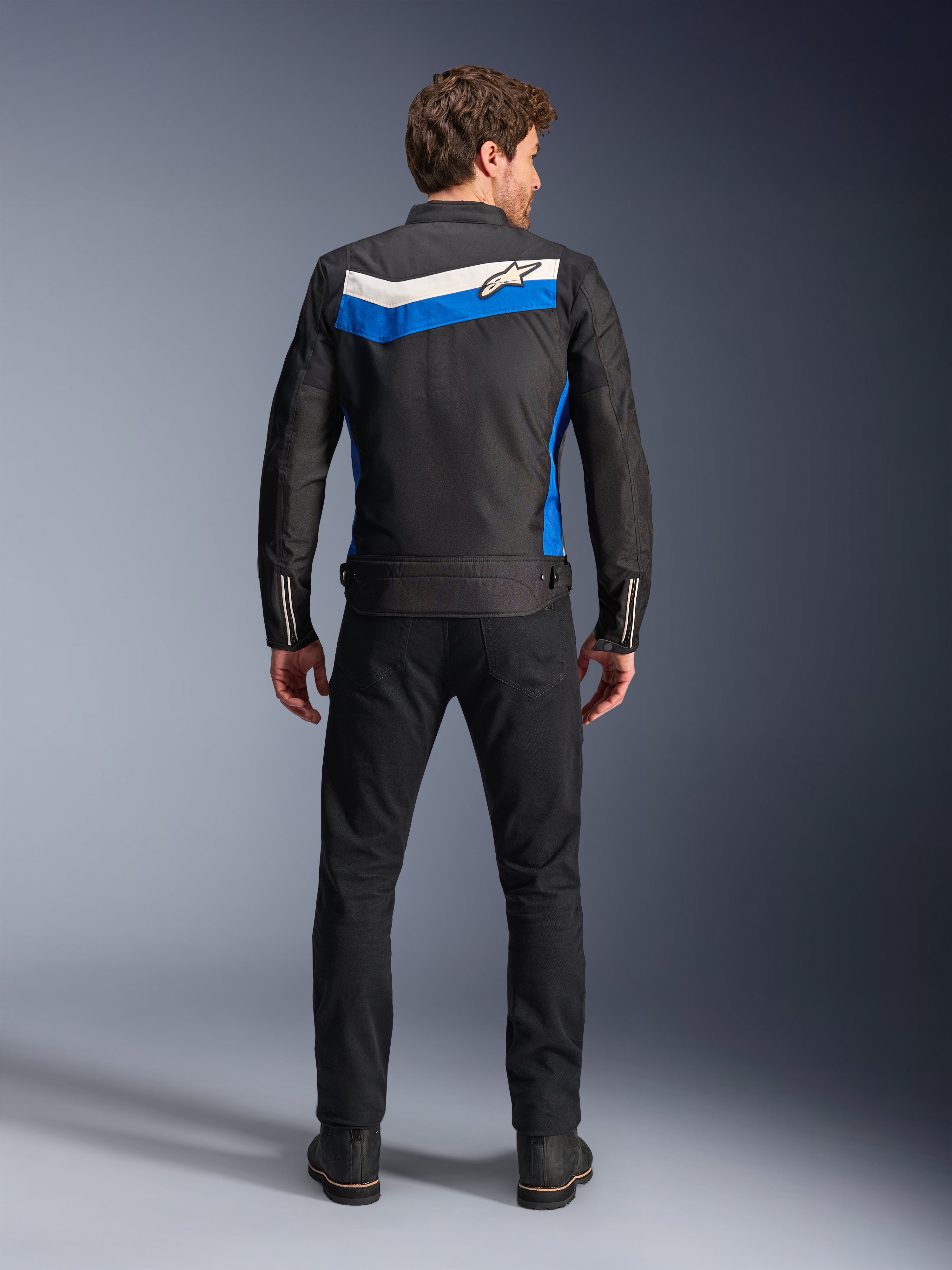 Alpinestars T-Dyno WR Jacket, motorcykeljacka i textil, färgkombinationen Black Blue Ecru, hel ryggvy på modell, med ett blått och off-white chevron-format band med Alpinestars-logotyp, kombinerad med svarta byxor och stövlar, vintageinspirerad urban mc-utrustning