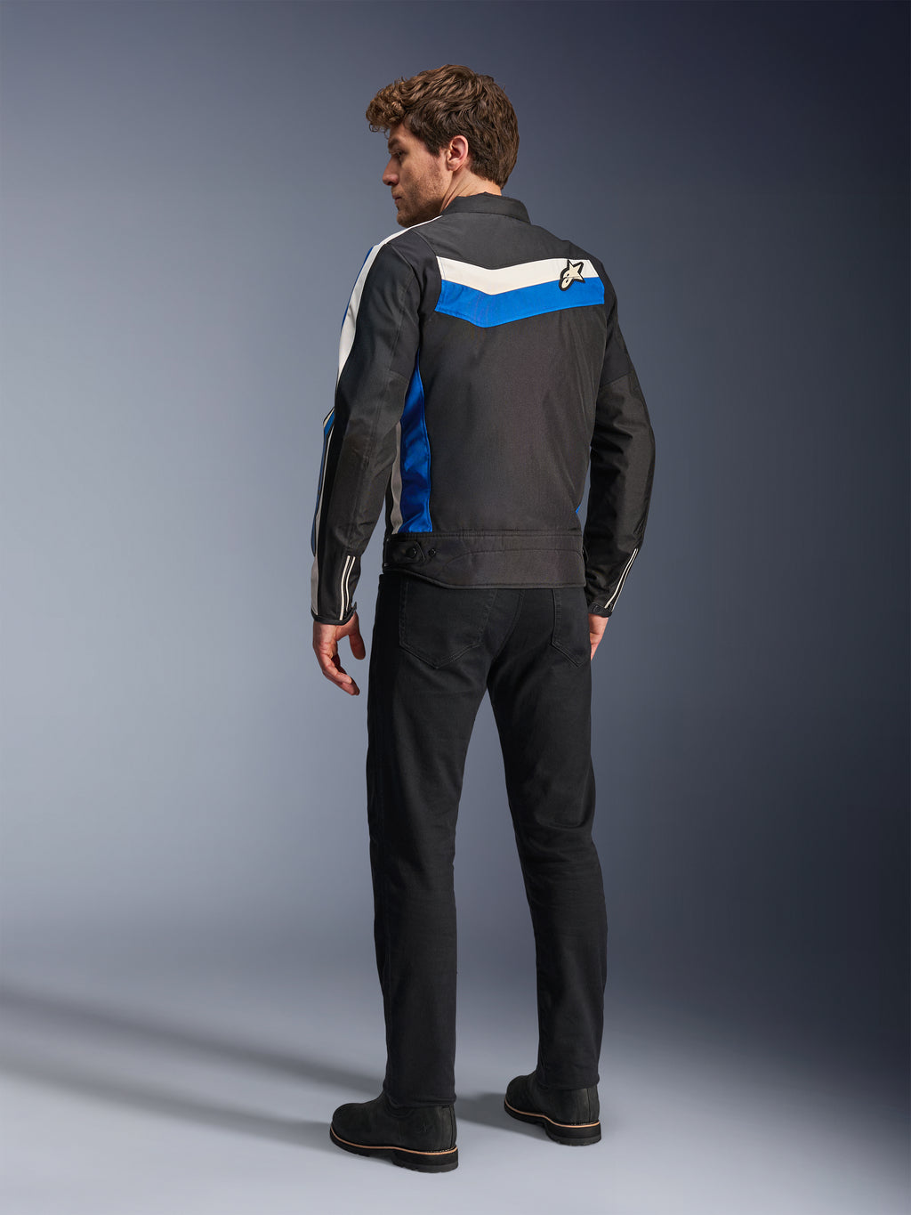 Alpinestars T-Dyno WR-jacka i Black Blue Ecru, sedd bakifrån på en manlig modell, med en vit och blå chevron-rand över axlarna och en stjärnlogotyp, vintageinspirerad textilmotorcykeljacka, bärs med svarta byxor och stövlar
