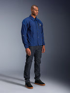 Alpinestars Revved WR-jacka, blå motorcykeljacka, bärs av en manlig modell med mörka denimjeans och svarta sneakers, med en vattenavvisande coachjacka-silhuett, tryckknappar framtill och en minimalistisk logotyp på bröstet