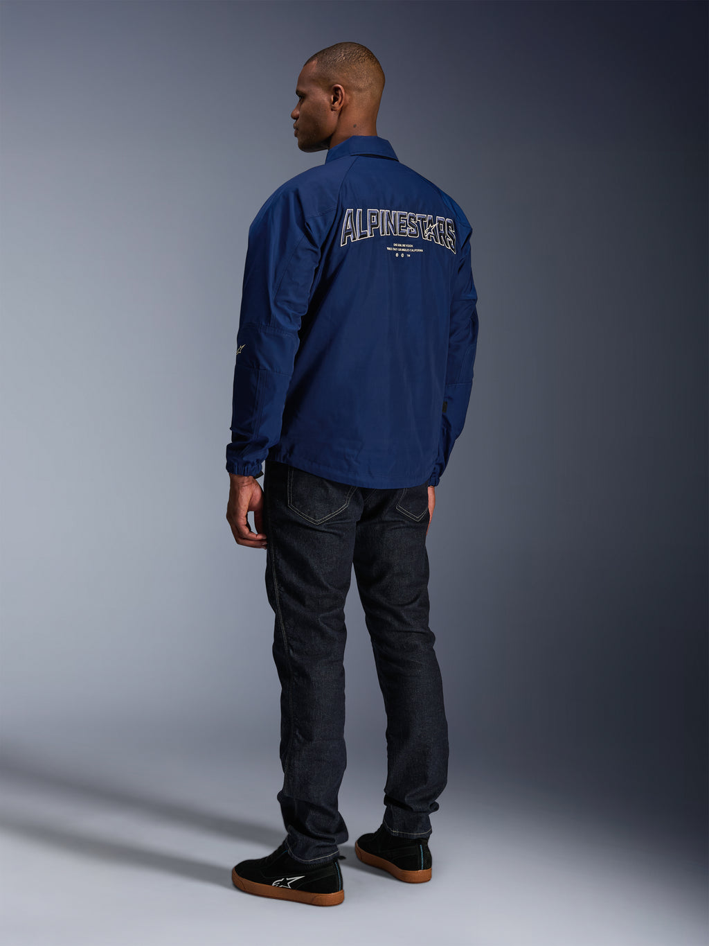 Modell som bär Alpinestars Revved WR-jacka i marinblått sedd bakifrån, med stor vit konturbroderad 'ALPINESTARS'-text över ryggen, matchad med mörka denimsbyxor och svarta sneakers, urban motorcykelestetik.