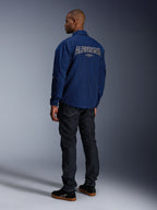 Modell som bär Alpinestars Revved WR-jacka i marinblått sedd bakifrån, med stor vit konturbroderad 'ALPINESTARS'-text över ryggen, matchad med mörka denimsbyxor och svarta sneakers, urban motorcykelestetik.