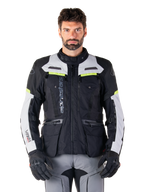 Man som bär Alpinestars Bogotá Pro Drystar®-jacka i färgerna svart, isgrå och neongul, allväders motorcykeljacka för adventure-touring, vattentät design med flera fickor och vertikal logotyp, matchad med svarta handskar och grå körbyxor