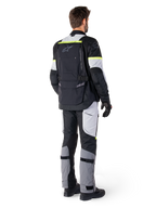 Bakifrån på en förare som bär Alpinestars Bogotá Pro Drystar®-jacka i Black Ice Gray Yellow Fluo, kombinerad med matchande adventure touring-byxor, med en stor ryggficka och gula fluorescerande detaljer, textilmotorcykelutrustning för alla säsonger.