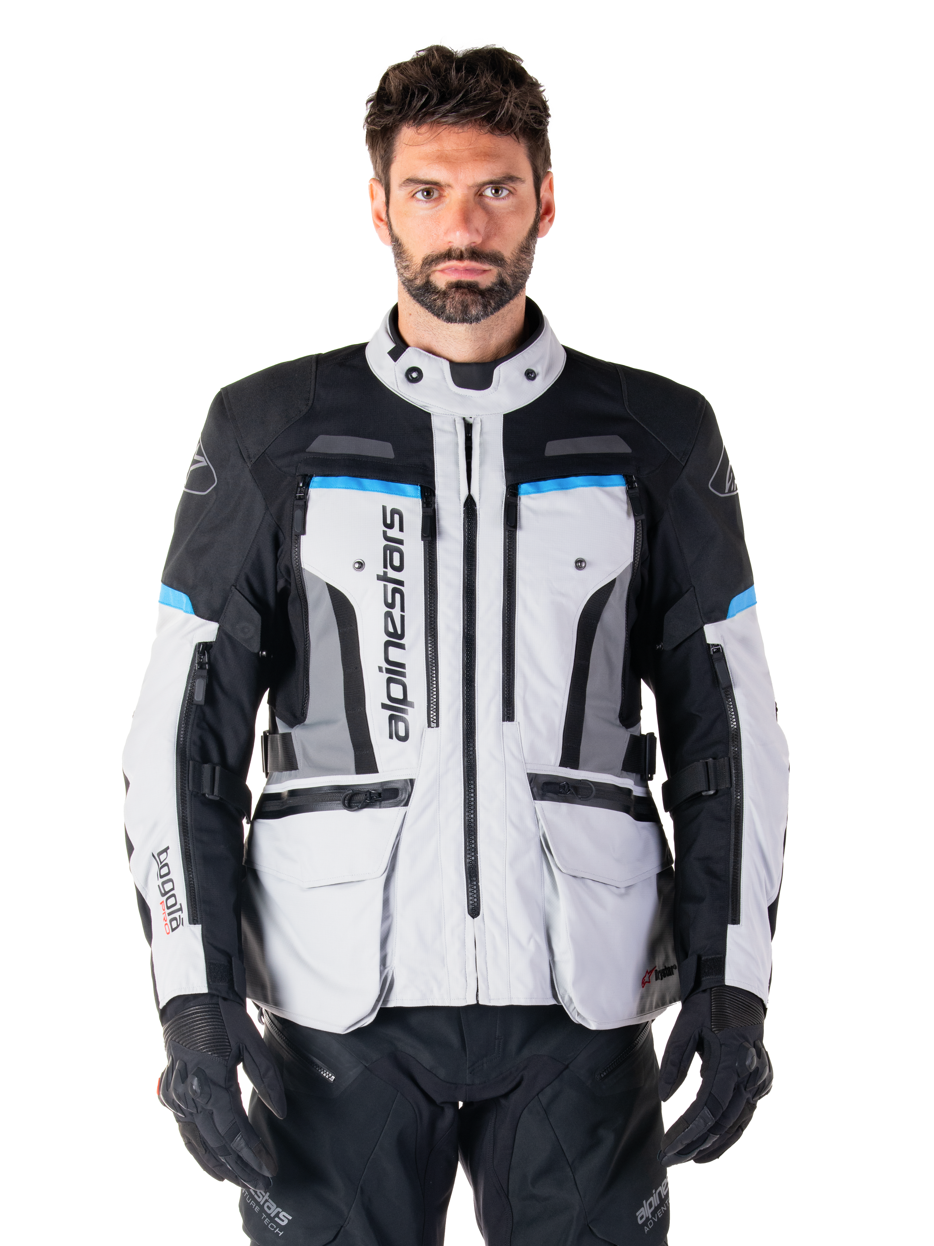 Alpinestars Bogota Pro Drystar-jacka, Adventure Touring-motorcykeljacka, isgrå med svarta och cyanblå detaljer, vy framifrån som visar ventilationspaneler och cargofickor, bärs av en modell med svarta mc-byxor och handskar