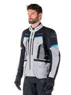 Alpinestars Bogota' Pro Drystar®-jacka, Ice Gray, svart och cyan, adventure-motorcykeljacka som bärs av en man med svarta handskar, teknisk design med flera fickor och ventilationspaneler, högpresterande körutrustning