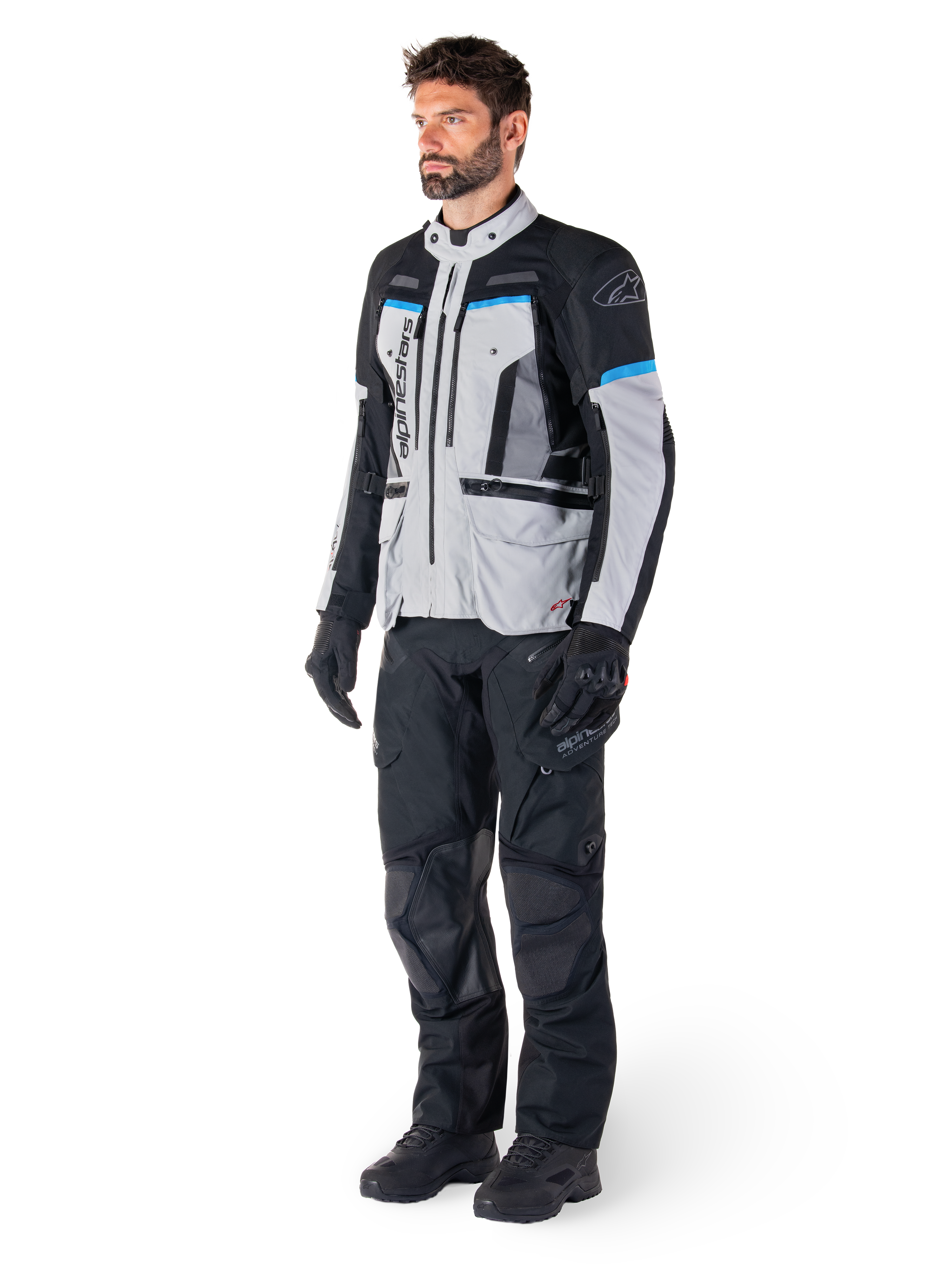 Manlig förare i Alpinestars Bogotá Pro Drystar®-jacka i Ice Gray, svart och cyan, matchad med svarta tekniska körbyxor och handskar, äventyrstouringutrustning för prestanda i alla väder