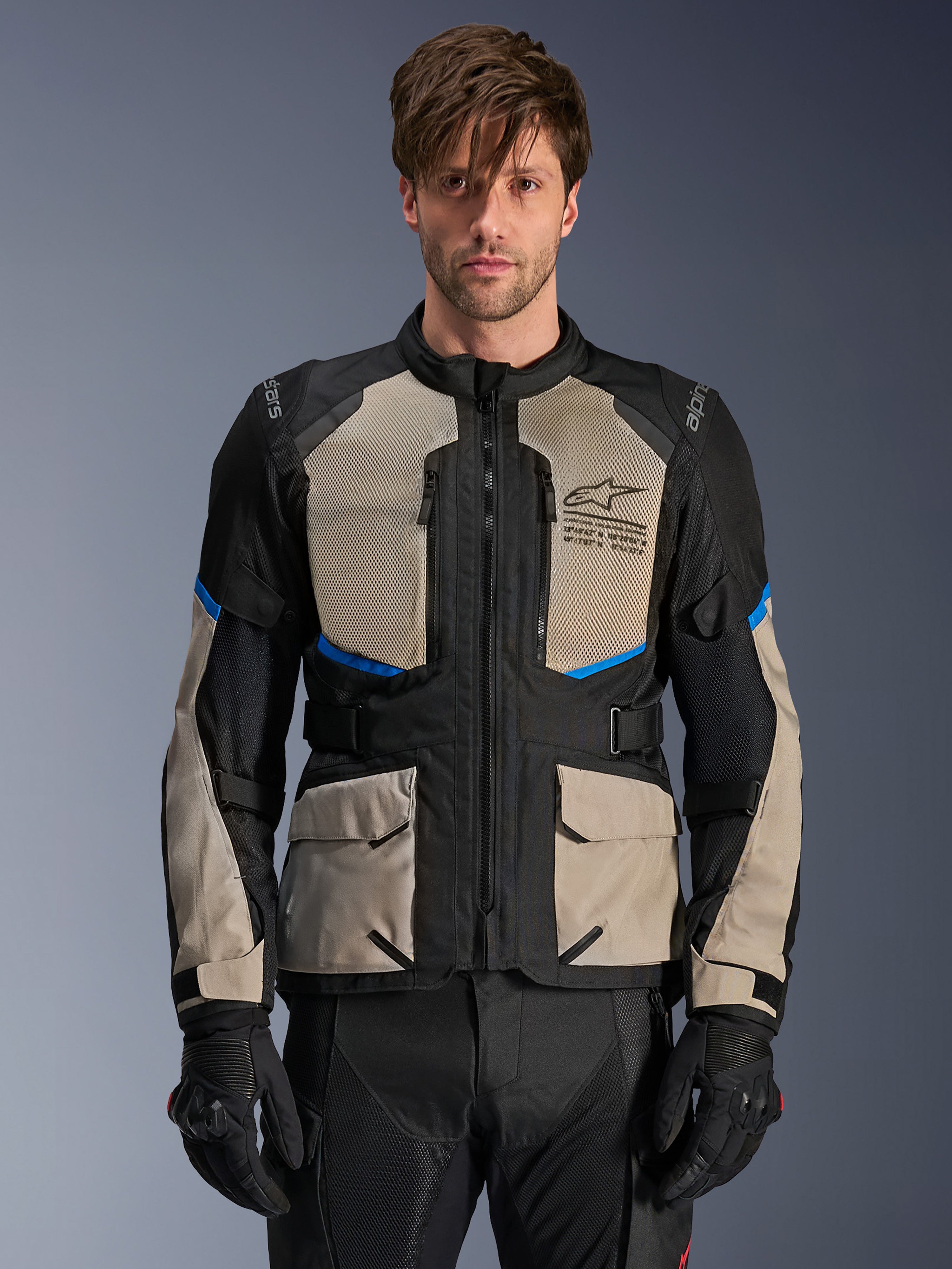 Alpinestars Andes Air Drystar jacka, adventure touring-jacka, Black Laurel Gray och Bright Blue, bärs av en modell med svarta handskar, stora mesh-paneler på bröstet för luftflöde, cargo-fickor fram, blå detaljer, skyddande motorcykelutrustning för touring