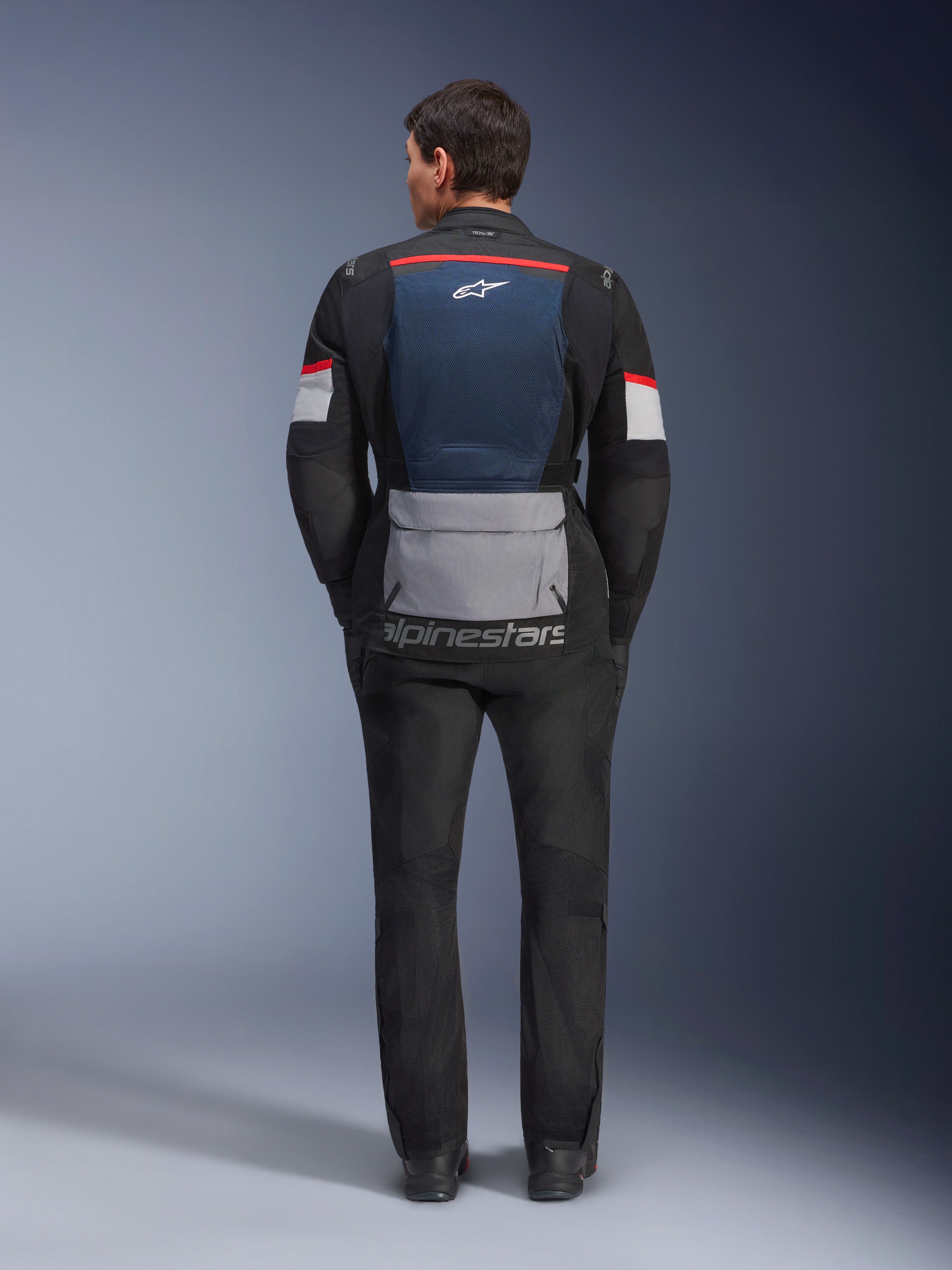 Alpinestars Andes Air Drystar-jacka, Deep Blue Black Ice Gray, vy bakifrån på modell, med blå mesh-panel på ryggen för ventilation, stor grå ficka nedtill, röda och vita detaljer på ärmarna, 'alpinestars' logotyp, kombinerad med svarta motorcykelbyxor för en komplett touring-outfit