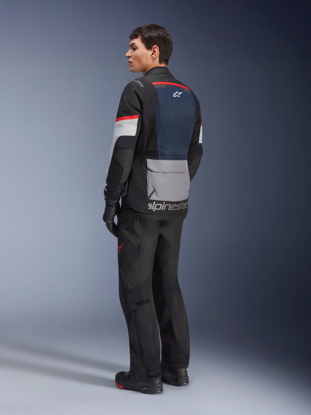 Manlig modell sedd snett bakifrån bärandes Alpinestars Andes Air Drystar®-jacka i Deep Blue Black Ice Gray, matchad med svarta motorcykelbyxor och handskar, med en blå ryggpanel i mesh, grå praktisk ficka på nedre delen av ryggen och röda detaljer på ärmarna, professionell touringutrustning för äventyrskörning.