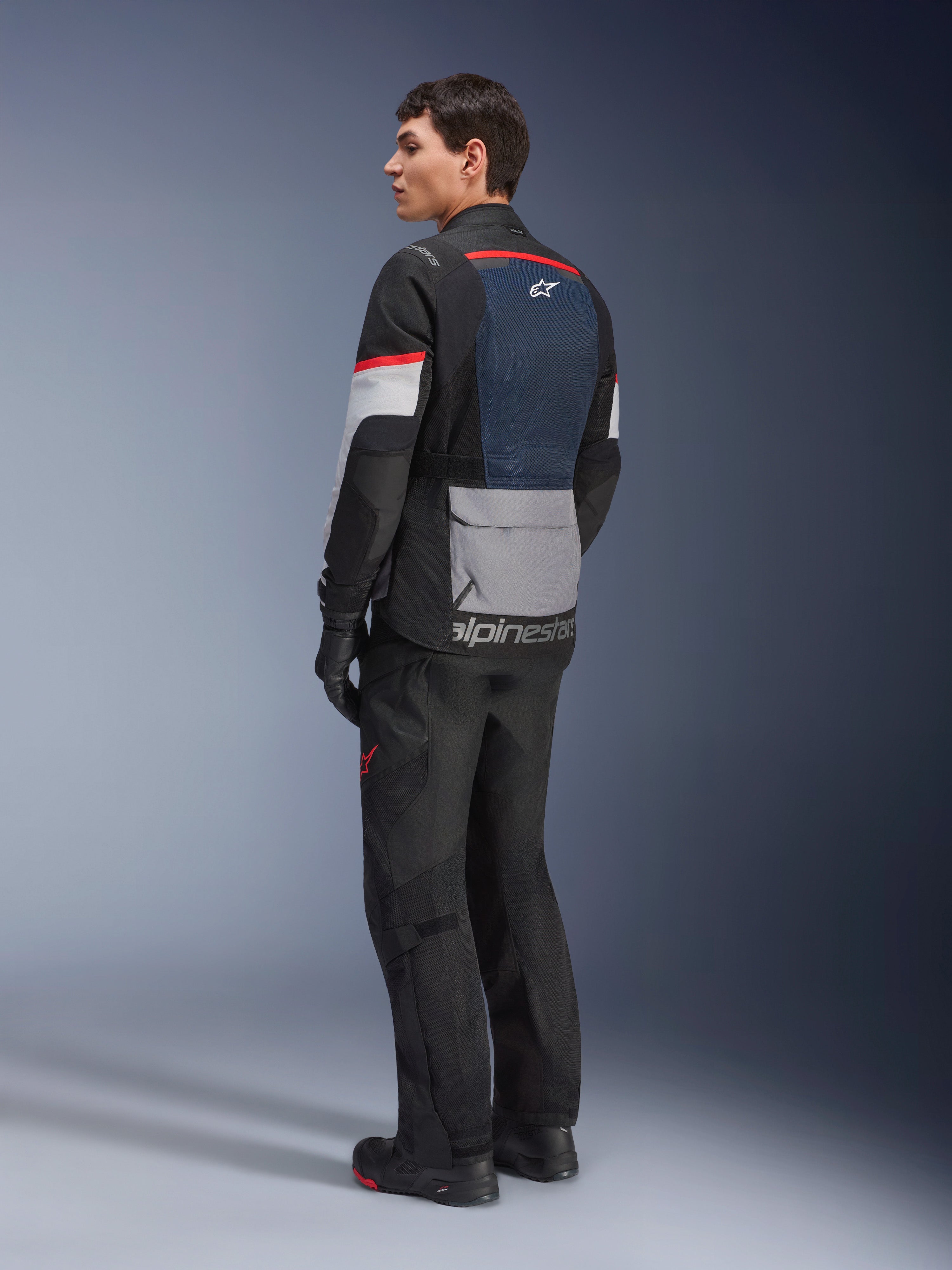 Manlig modell sedd snett bakifrån bärandes Alpinestars Andes Air Drystar®-jacka i Deep Blue Black Ice Gray, matchad med svarta motorcykelbyxor och handskar, med en blå ryggpanel i mesh, grå praktisk ficka på nedre delen av ryggen och röda detaljer på ärmarna, professionell touringutrustning för äventyrskörning.