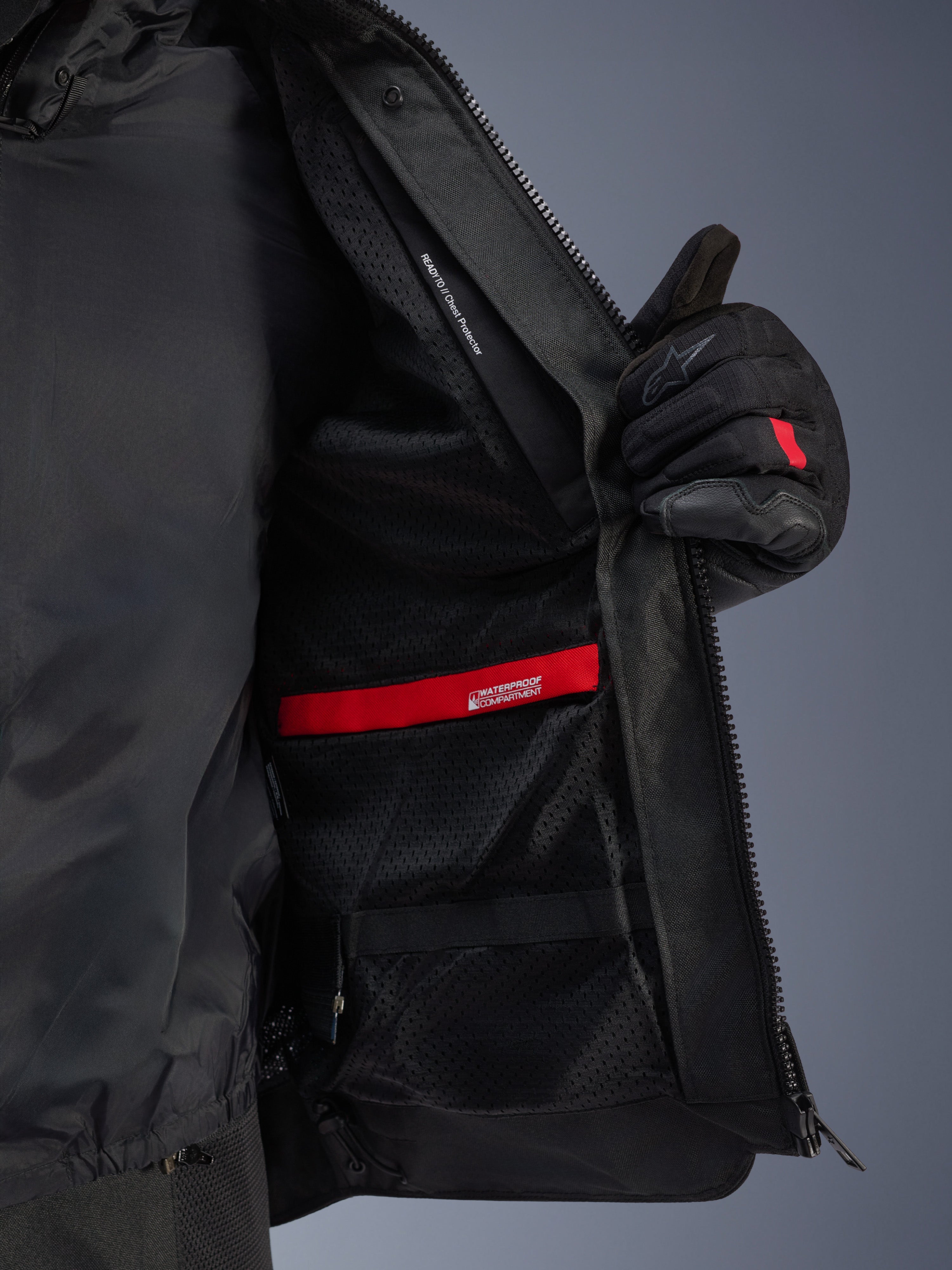 Detaljvy av insidan på Alpinestars Andes Air Drystar® Jacket, som framhäver det andningsbara svarta mesh-fodret, det röda vattentäta facket och fickan för bröstskydd, visas hållas öppen av en svart motorcykelhandske
