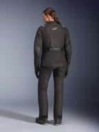 Kvinnlig förare sedd bakifrån klädd i Alpinestars Stella Andes V4 Drystar®-jacka, svart, kombinerad med matchande motorcykelbyxor, vit Astar-logotyp på ryggen, förlängd fåll baktill, komplett adventure-touring-outfit inklusive svarta handskar och stövlar