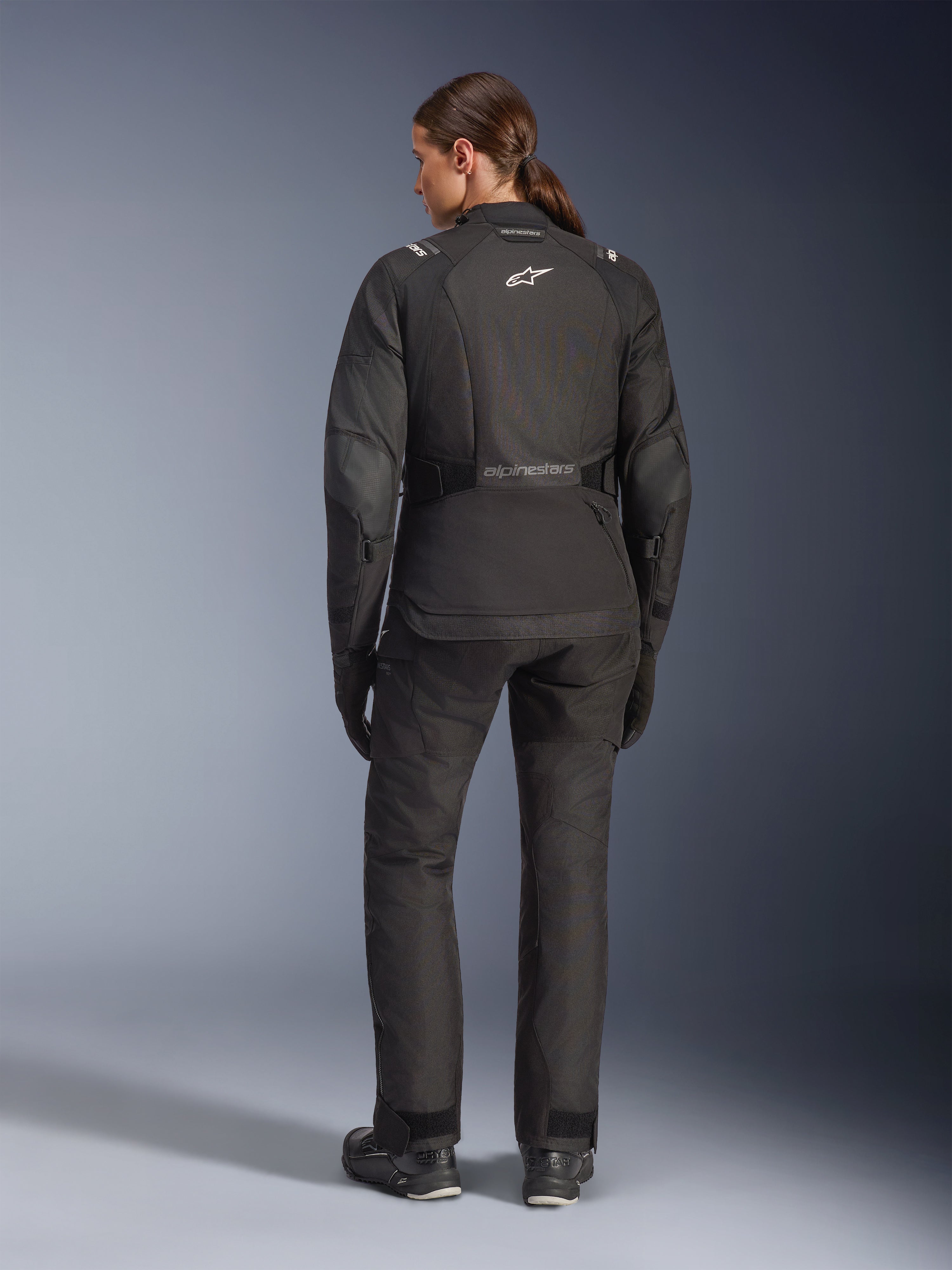 Kvinnlig förare sedd bakifrån klädd i Alpinestars Stella Andes V4 Drystar®-jacka, svart, kombinerad med matchande motorcykelbyxor, vit Astar-logotyp på ryggen, förlängd fåll baktill, komplett adventure-touring-outfit inklusive svarta handskar och stövlar