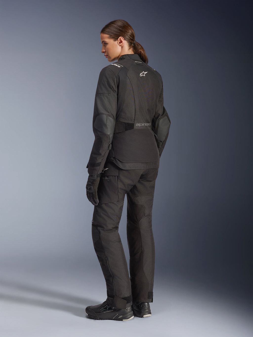 Kvinnlig förare i Alpinestars Stella Andes V4 Drystar®-jacka, svart motorcykeljacka för äventyrskörning, 3/4-vy bakifrån, matchad med tillhörande textilbyxor och handskar, vita Alpinestars-logotyper på ryggen och i midjan, förstärkta skyddspaneler och förlängd bakre fåll.
