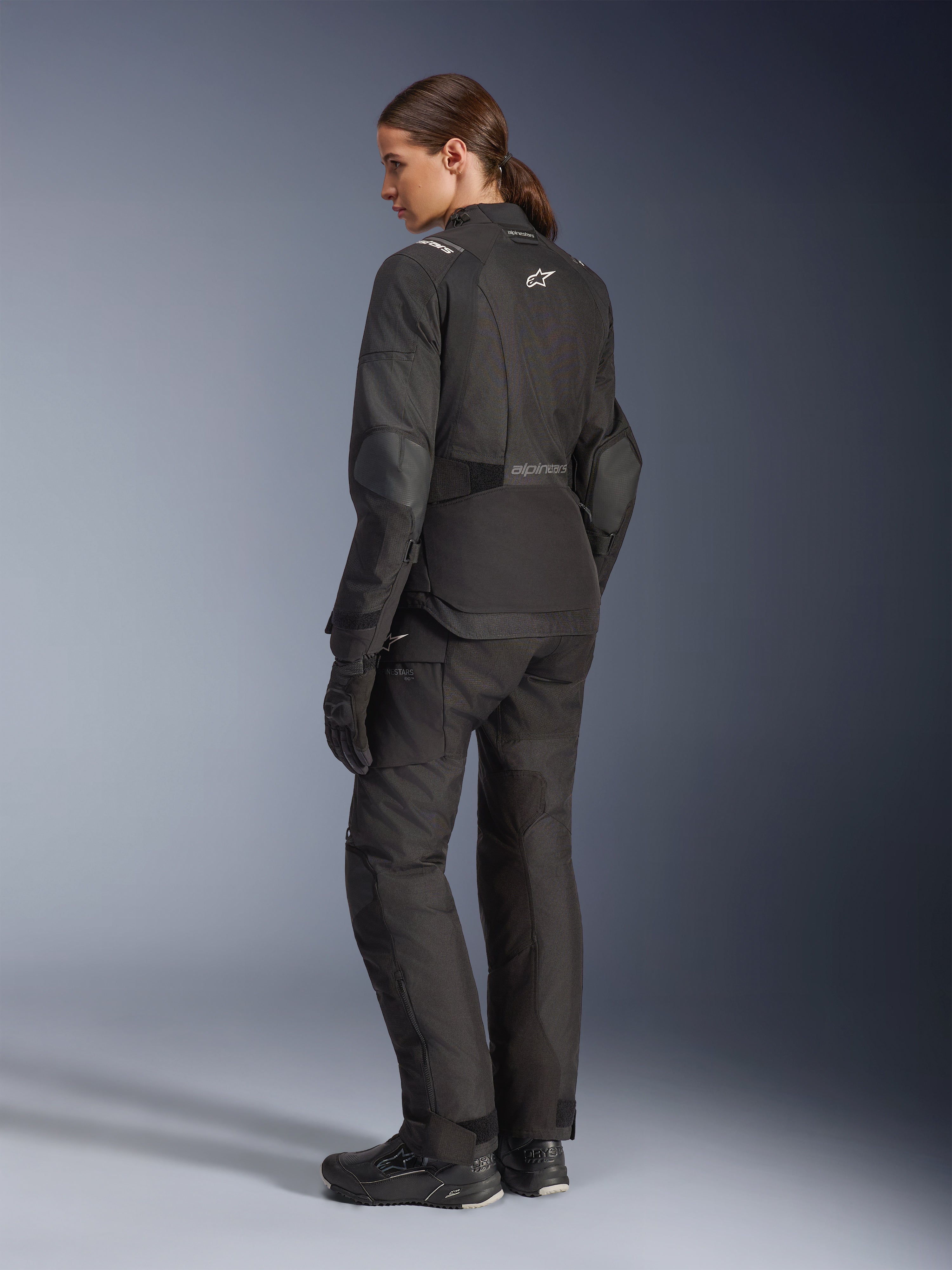 Kvinnlig förare i Alpinestars Stella Andes V4 Drystar®-jacka, svart motorcykeljacka för äventyrskörning, 3/4-vy bakifrån, matchad med tillhörande textilbyxor och handskar, vita Alpinestars-logotyper på ryggen och i midjan, förstärkta skyddspaneler och förlängd bakre fåll.