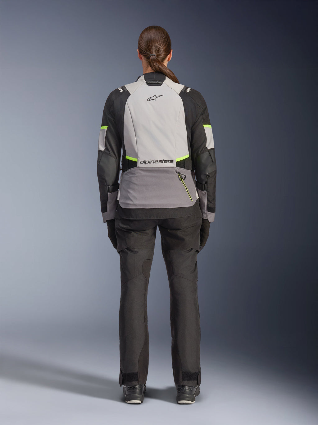 Kvinnlig förare sedd bakifrån iförd Alpinestars Stella Andes V4 Drystar®-jacka i isgrått, mörkgrått och svart med gula fluo-detaljer, utrustad med en stor bakre förvaringsficka och Alpinestars-logotyp, matchad med svarta adventure-byxor, mc-touringutrustning för alla väder