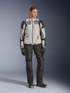 Kvinnlig modell bär Alpinestars Stella Andes V4 Drystar®-jacka i Ice Gray, Dark Gray och svart med Yellow Fluo-detaljer, kombinerat med svarta motorcykelbyxor och handskar, helkroppsbild i studiomiljö av äventyrsutrustning.