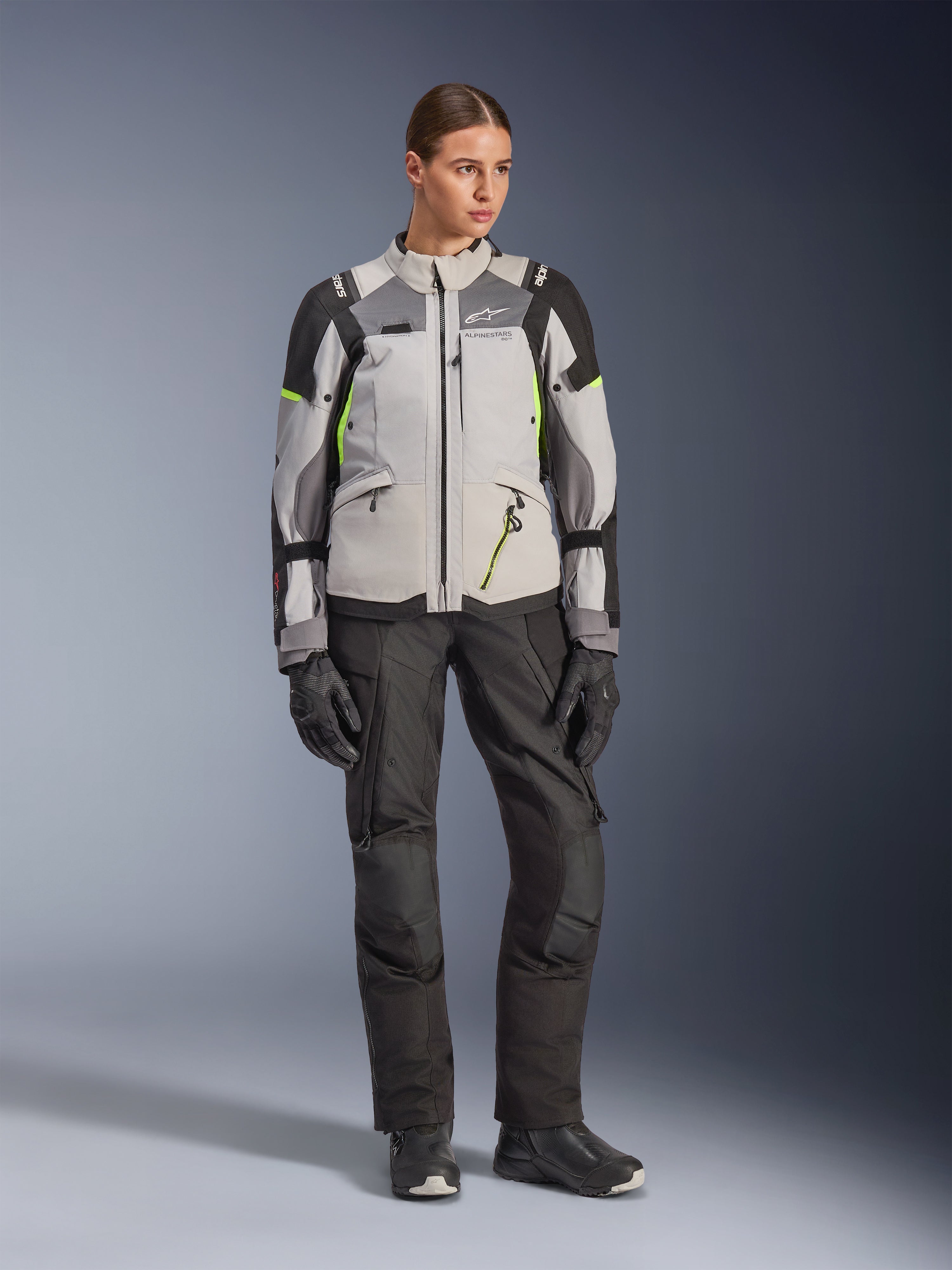 Kvinnlig modell bär Alpinestars Stella Andes V4 Drystar®-jacka i Ice Gray, Dark Gray och svart med Yellow Fluo-detaljer, kombinerat med svarta motorcykelbyxor och handskar, helkroppsbild i studiomiljö av äventyrsutrustning.