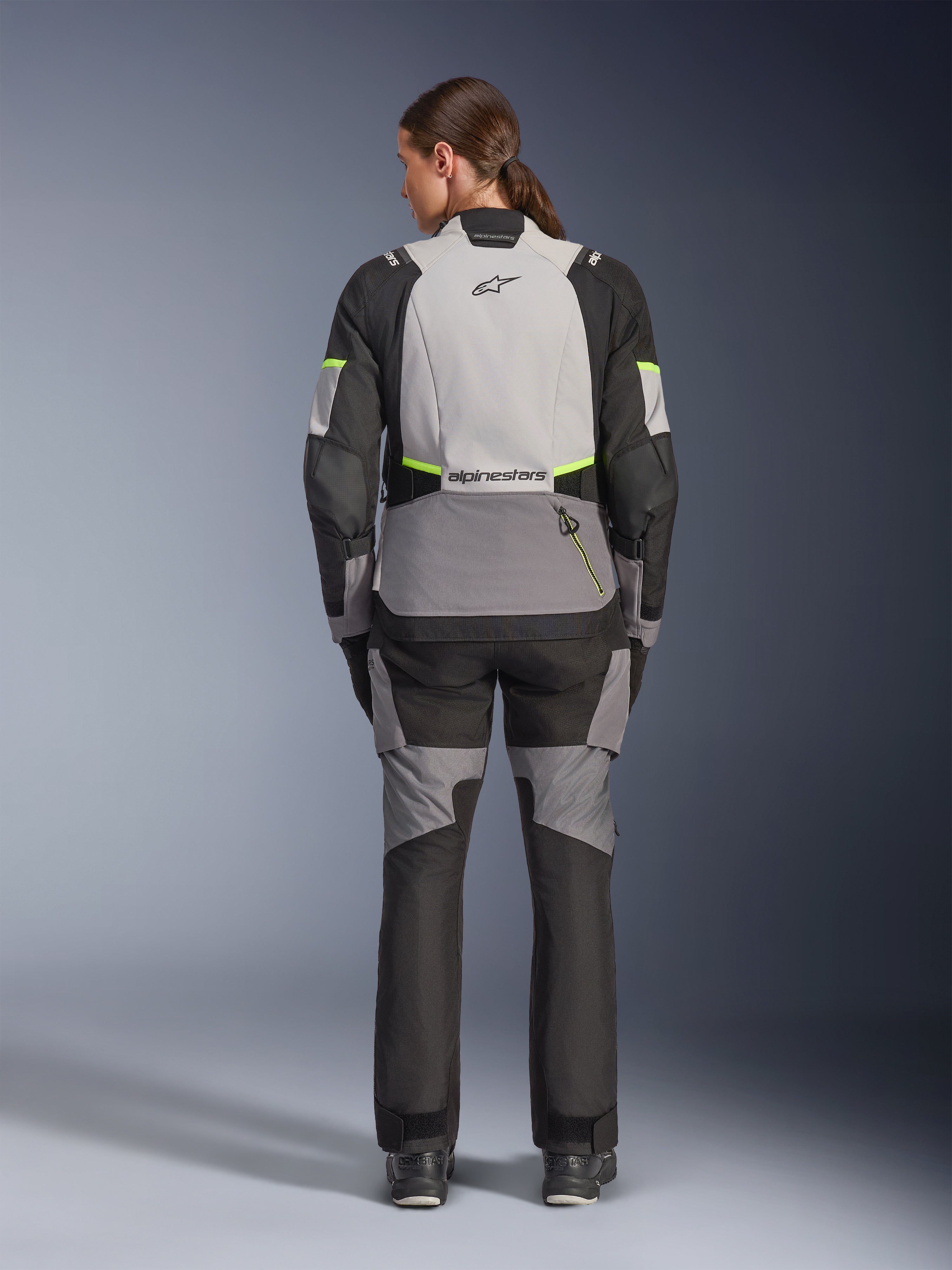 Kvinnlig förare sedd bakifrån i Alpinestars Stella Andes V4 Drystar®-jacka och matchande byxor, Ice Gray och Dark Gray med detaljer i Black och Yellow Fluo, adventure-touring motorcykelutrustning med hållbar textilkonstruktion och reflekterande logotyper