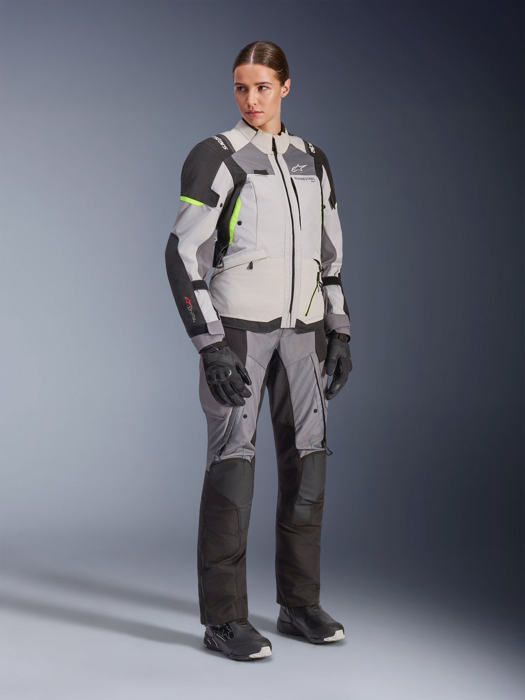 Kvinna i Alpinestars Stella Andes V4 Drystar®-jacka och matchande byxor, Ice Gray och Dark Gray med Yellow Fluo-detaljer, komplett äventyrsställ med svarta handskar och stövlar, helfigursbild i studio