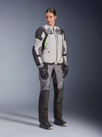 Kvinna i Alpinestars Stella Andes V4 Drystar®-jacka och matchande byxor, Ice Gray och Dark Gray med Yellow Fluo-detaljer, komplett äventyrsställ med svarta handskar och stövlar, helfigursbild i studio
