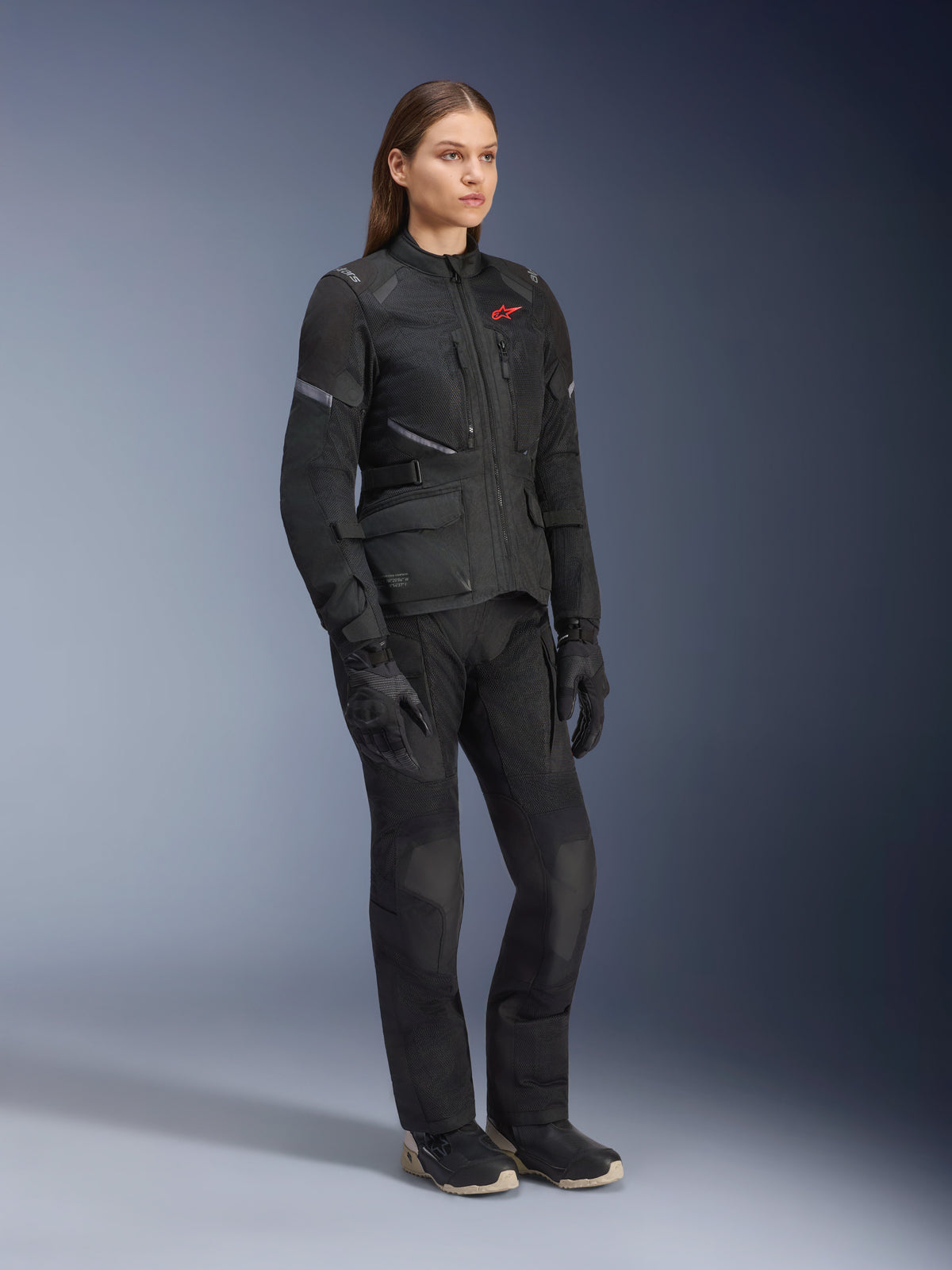 Kvinna i Alpinestars Stella Andes Air Drystar®-jacka, svart textiljacka för adventure touring, buren med matchande svarta byxor och handskar, med stora ventilationspaneler i mesh och en röd logotyp, koordinerad outfit för motorcykelkörning för damer