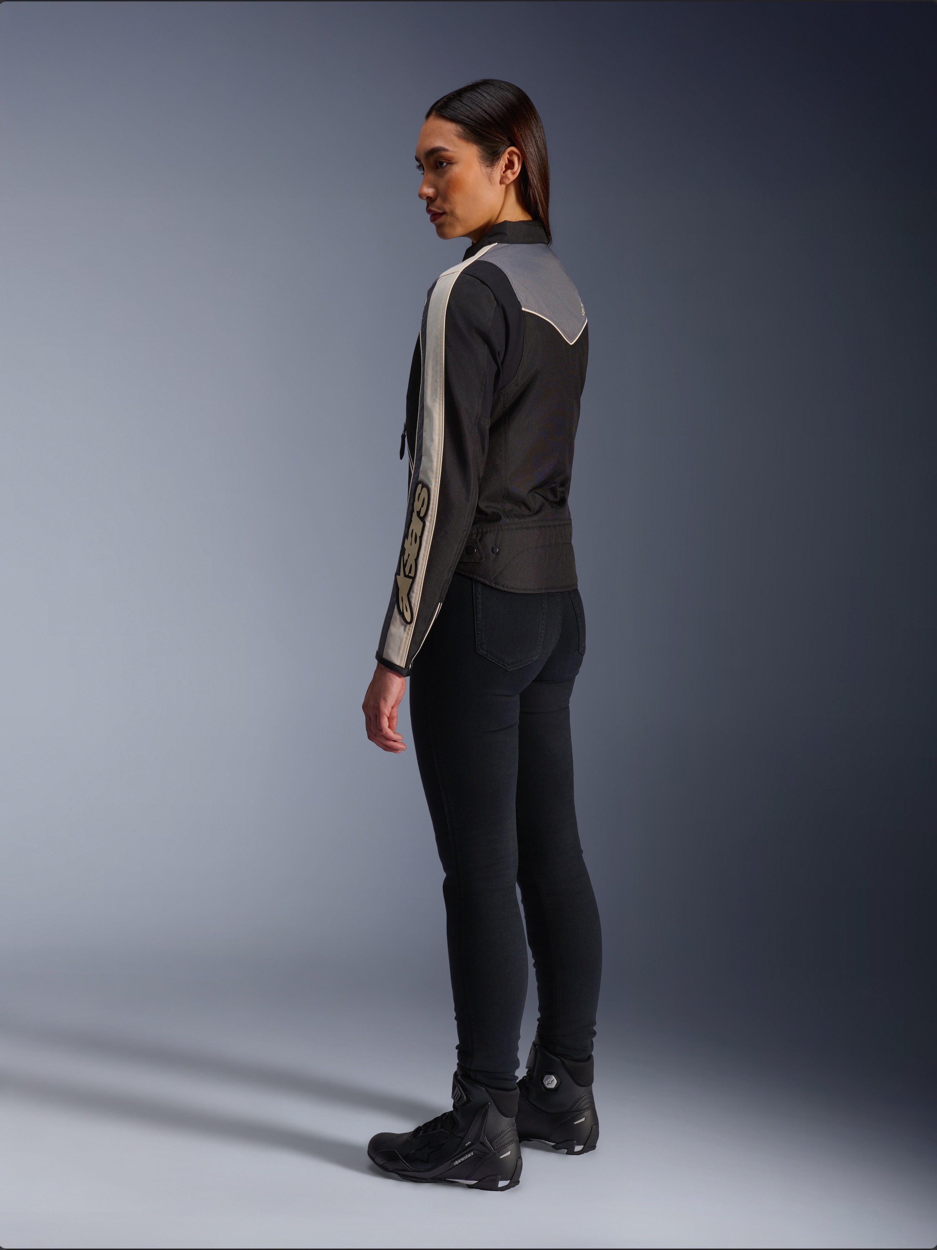 Female model viewed from the side and back wearing the Alpinestars Stella T-Dyno WR Jacket in Black Dark Gray Ecru, featuring vintage-inspired design with ecru sleeve stripes and a gray back panel, worn with black pants and black riding shoes.