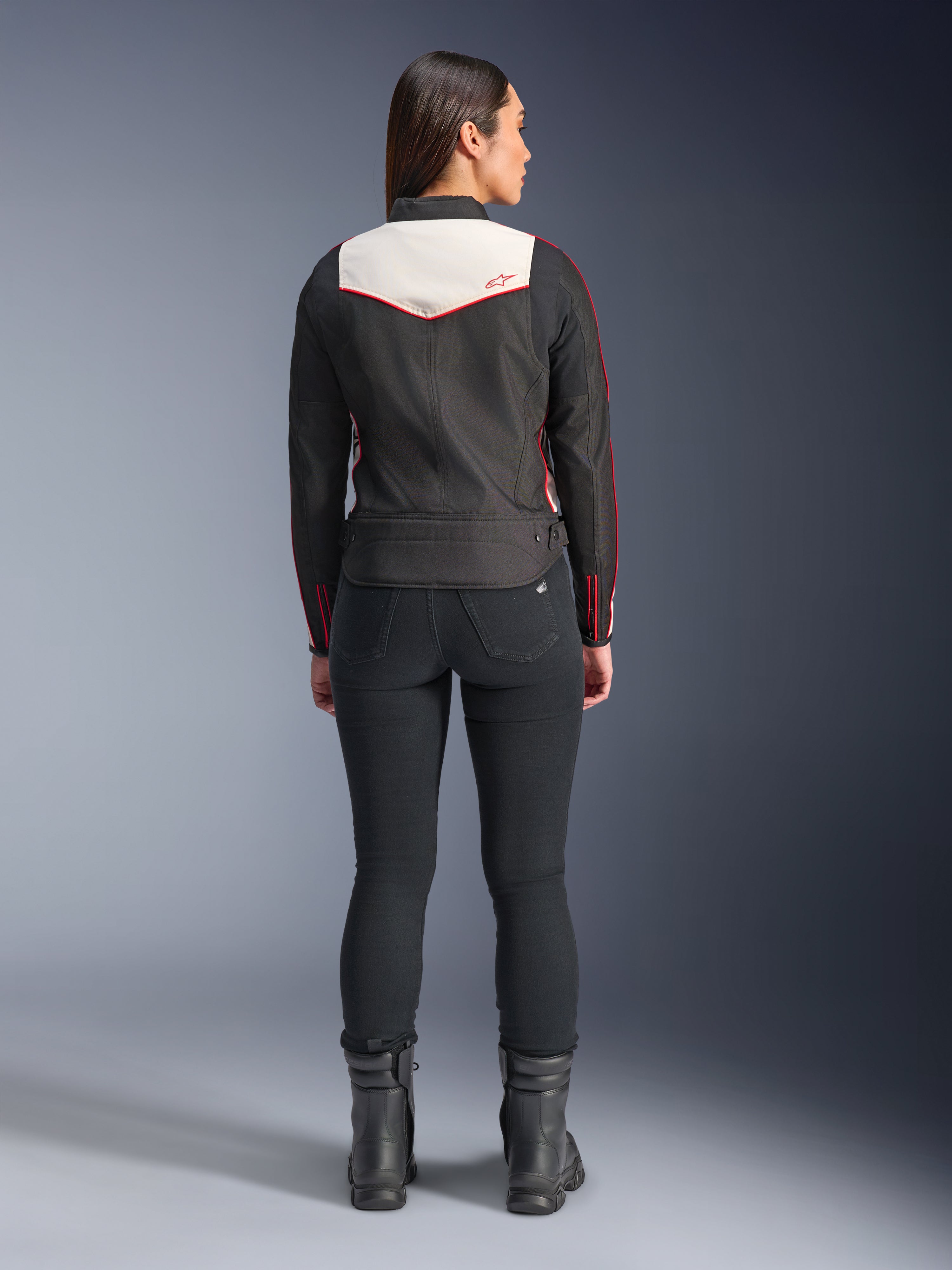 Rear view of a female model wearing the Alpinestars Stella T-Dyno WR Jacket in Black Ecru Bright Red, featuring a prominent cream back panel and red logo accents, styled with black pants and boots, a vintage-inspired textile motorcycle jacket.