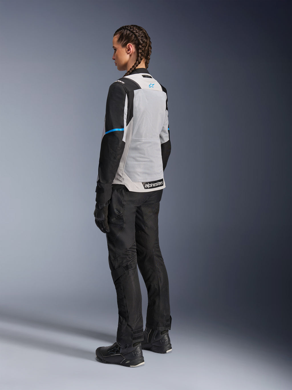 Female rider wearing the Alpinestars Stella ST-2 Air Jacket in Ice Gray and Black, rear three-quarter view highlighting the large light gray mesh back panel, black sleeves with a blue accent stripe, and a blue Alpinestars logo, paired with black riding pants, gloves, and boots for a complete adventure touring outfit