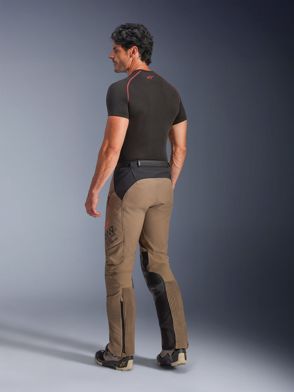 Alpinestars AMT-8 Stretch Drystar® XF-byxor i Walnut Black, äventyrstouring-motorcykelbyxor sedda från en trekvartsvy bakifrån, valnötsbrun textil med svarta paneler på sätet och insidan av benen, dragspelsstretchpaneler på vaderna, AMT-logotyp på sidan, bärs med ett svart underställ.