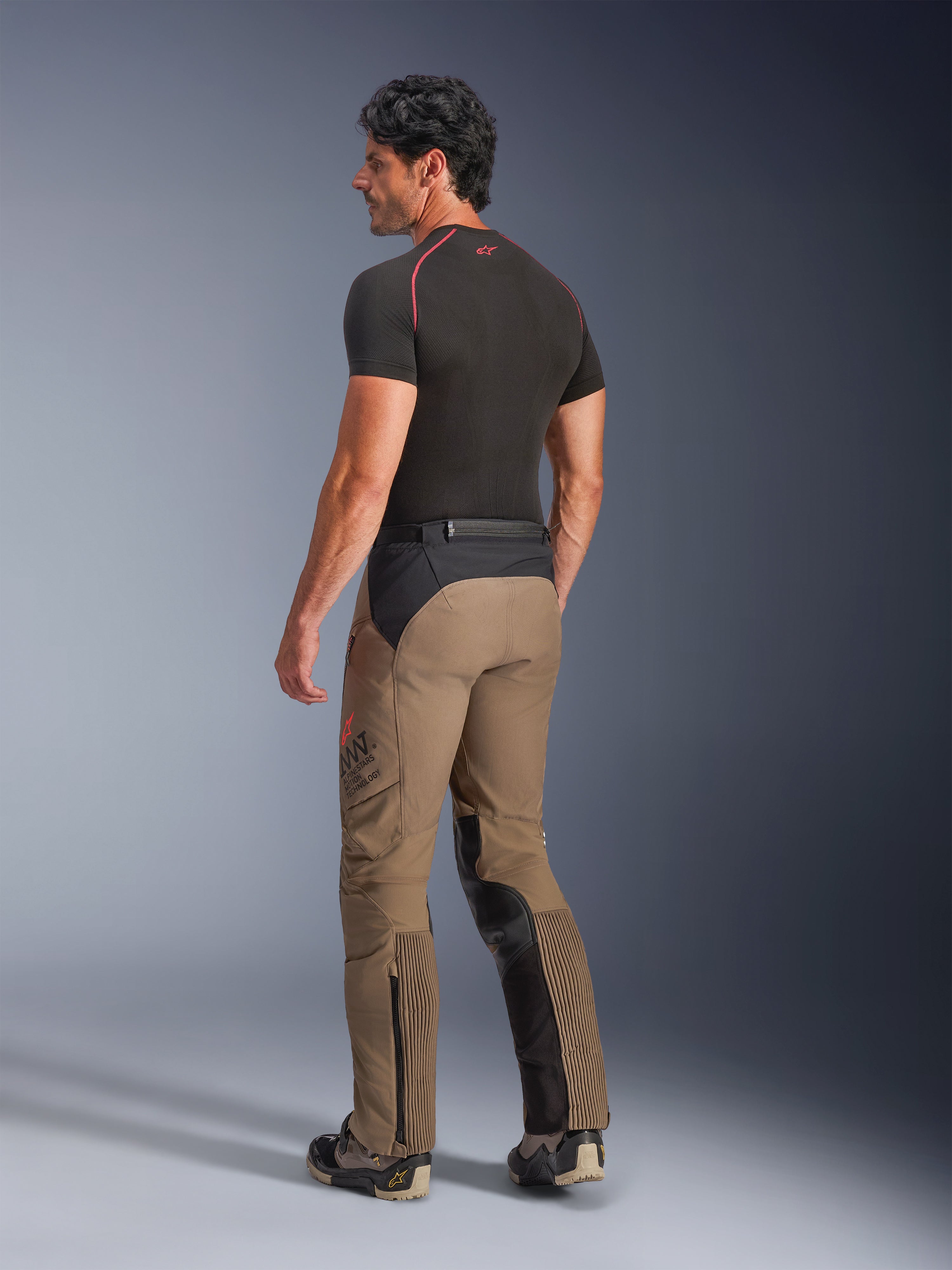 Alpinestars AMT-8 Stretch Drystar® XF-byxor i Walnut Black, äventyrstouring-motorcykelbyxor sedda från en trekvartsvy bakifrån, valnötsbrun textil med svarta paneler på sätet och insidan av benen, dragspelsstretchpaneler på vaderna, AMT-logotyp på sidan, bärs med ett svart underställ.