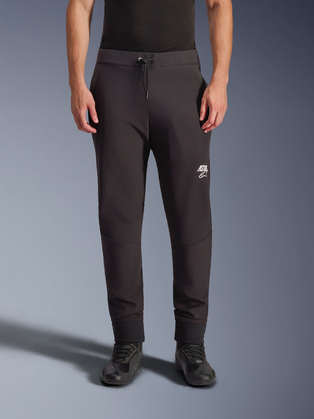 Alpinestars Aeroshell Pants, Black softshell motorcycle pants, jogger-style athletic fit with drawstring waist and elastic cuffs, featuring a white Astars logo, worn with black riding shoes, modern crossover gear for road and casual use