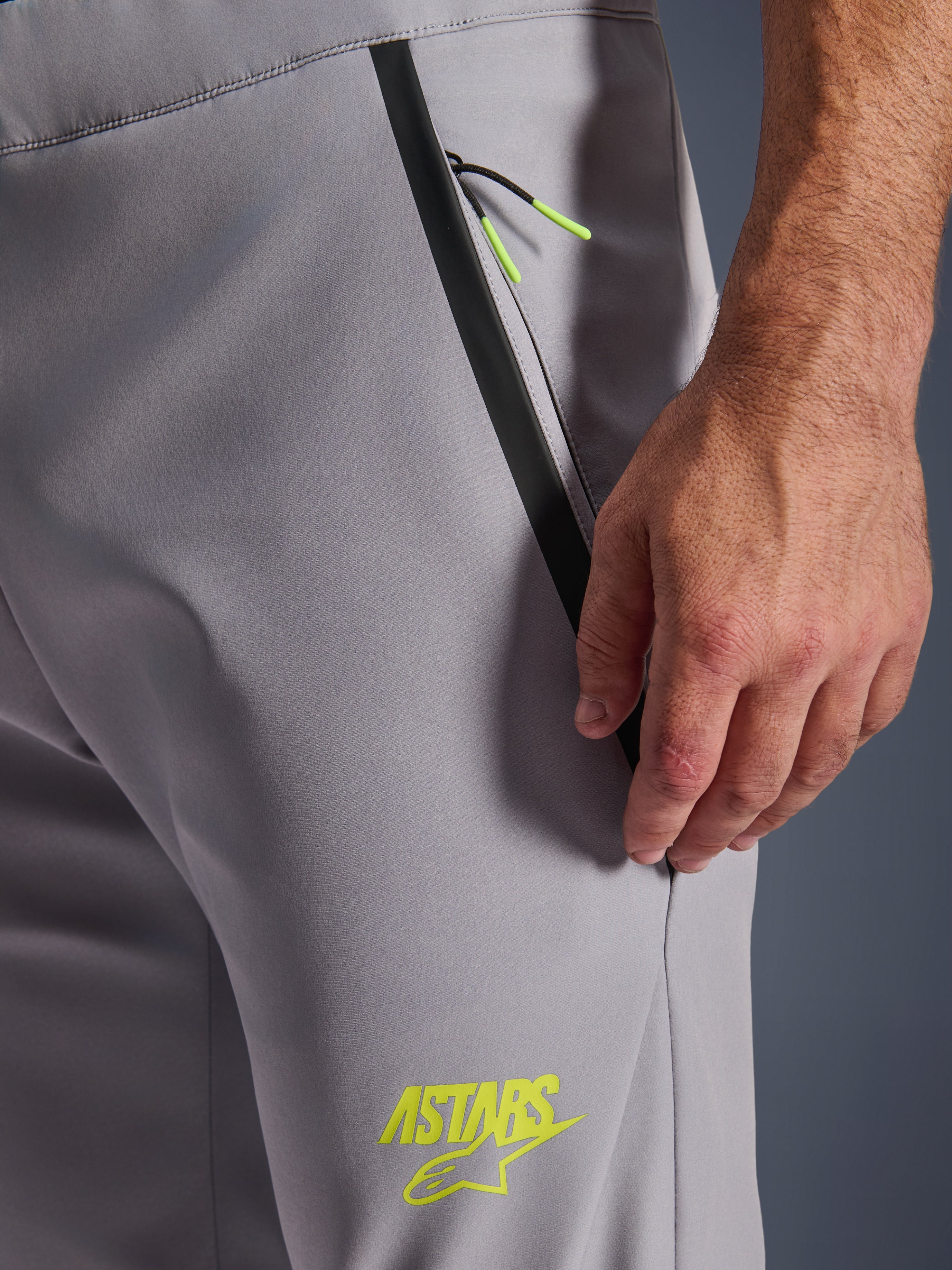 Alpinestars Aeroshell-byxor, mellangrå med neongula detaljer, närbild av framfickan med dragkedja, svart tejpad kant och klargula dragsnören, gul ASTARS-logotyp på låret, stretchigt softshell-material för motorcykelkörning.