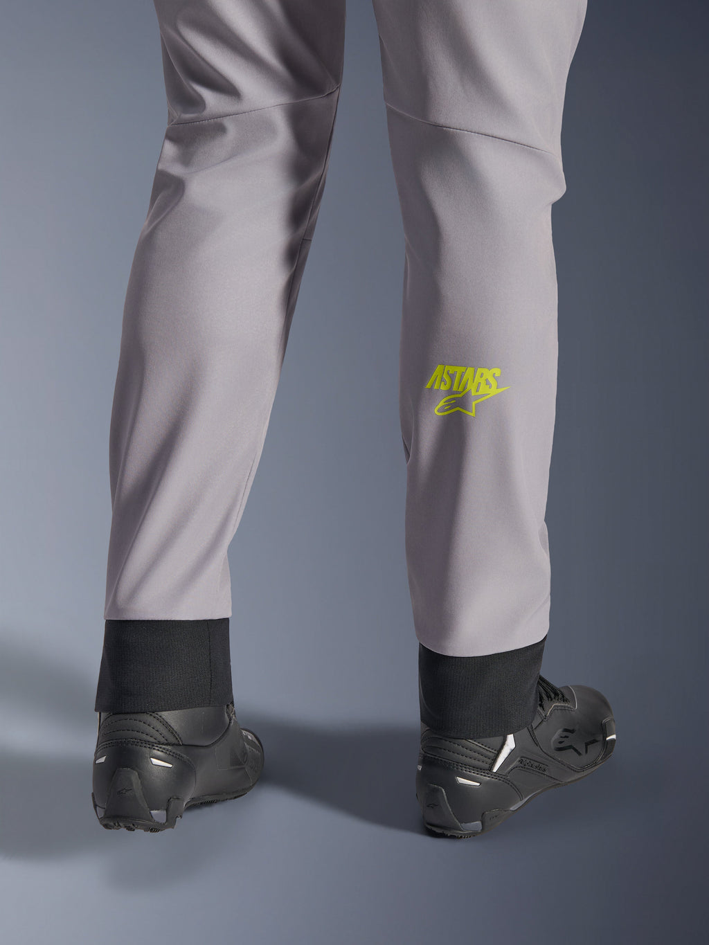 Alpinestars Aeroshell byxor, mellangrå med neongul Astars-logotyp på vaden, sedda bakifrån med svarta motorcykelskor, med en elastisk nedre fåll och en modern avsmalnande silhuett