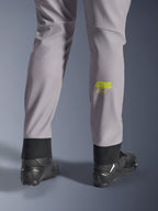 Alpinestars Aeroshell byxor, mellangrå med neongul Astars-logotyp på vaden, sedda bakifrån med svarta motorcykelskor, med en elastisk nedre fåll och en modern avsmalnande silhuett