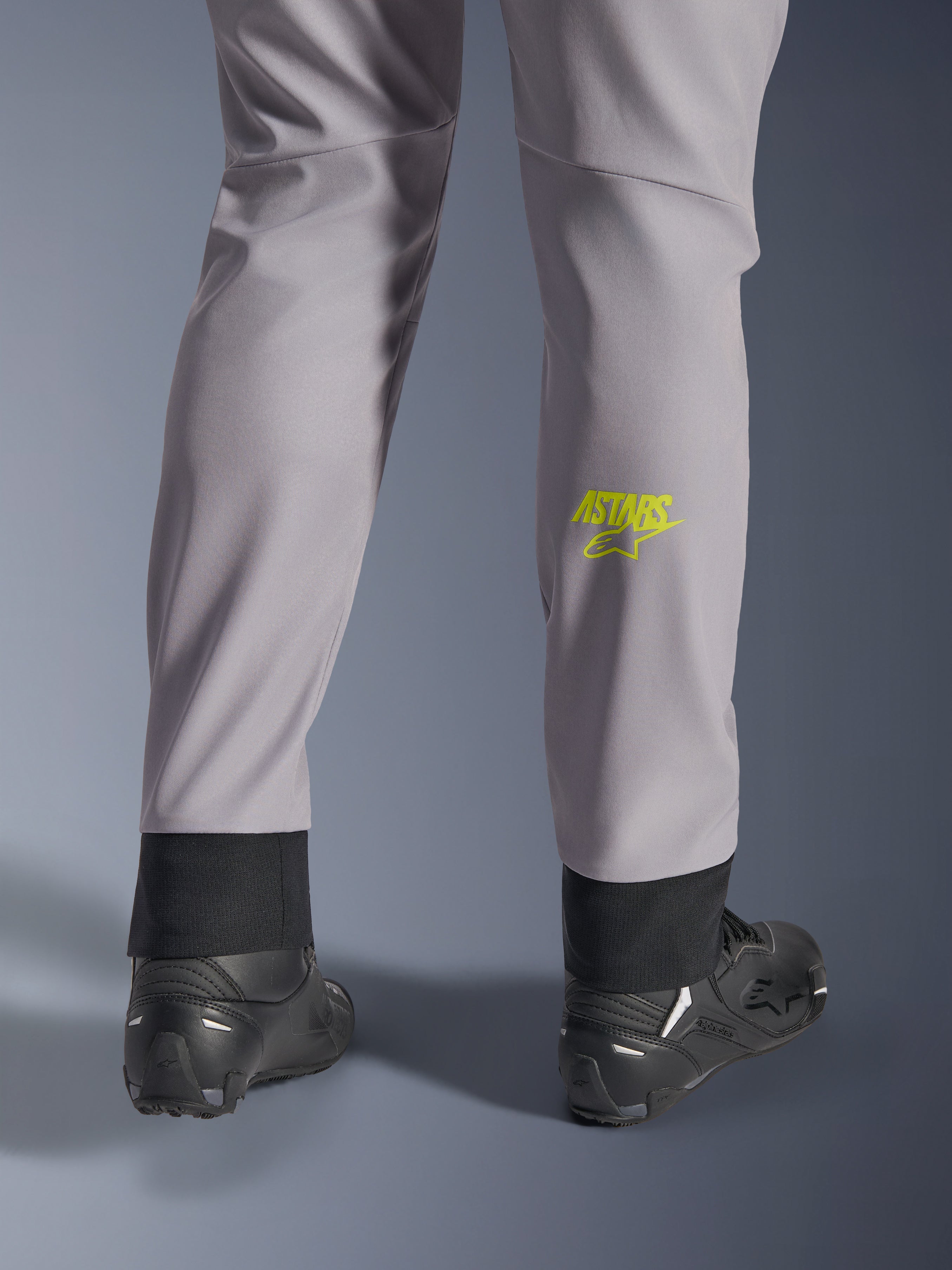 Alpinestars Aeroshell byxor, mellangrå med neongul Astars-logotyp på vaden, sedda bakifrån med svarta motorcykelskor, med en elastisk nedre fåll och en modern avsmalnande silhuett