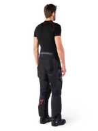 Alpinestars Hyde XT Drystar® Pants, Adventure Touring Pants, Black and Bright Red, rear view of a rider wearing the pants with a black base layer and black boots, featuring durable textile construction with a red star logo on the lower leg and gray Alpinestars branding on the waistband