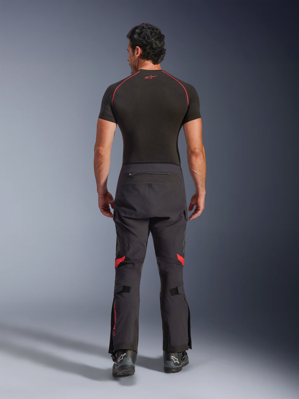 Alpinestars Halo Pro Drystar® XF laminerade byxor, äventyrsmotorcykelbyxor, Black Dark Shadow med röda detaljer, sedda bakifrån bärs med ett svart kortärmat underställ och svarta äventyrsstövlar, visar en hållbar 3-lagers laminerad konstruktion och ergonomisk passform.