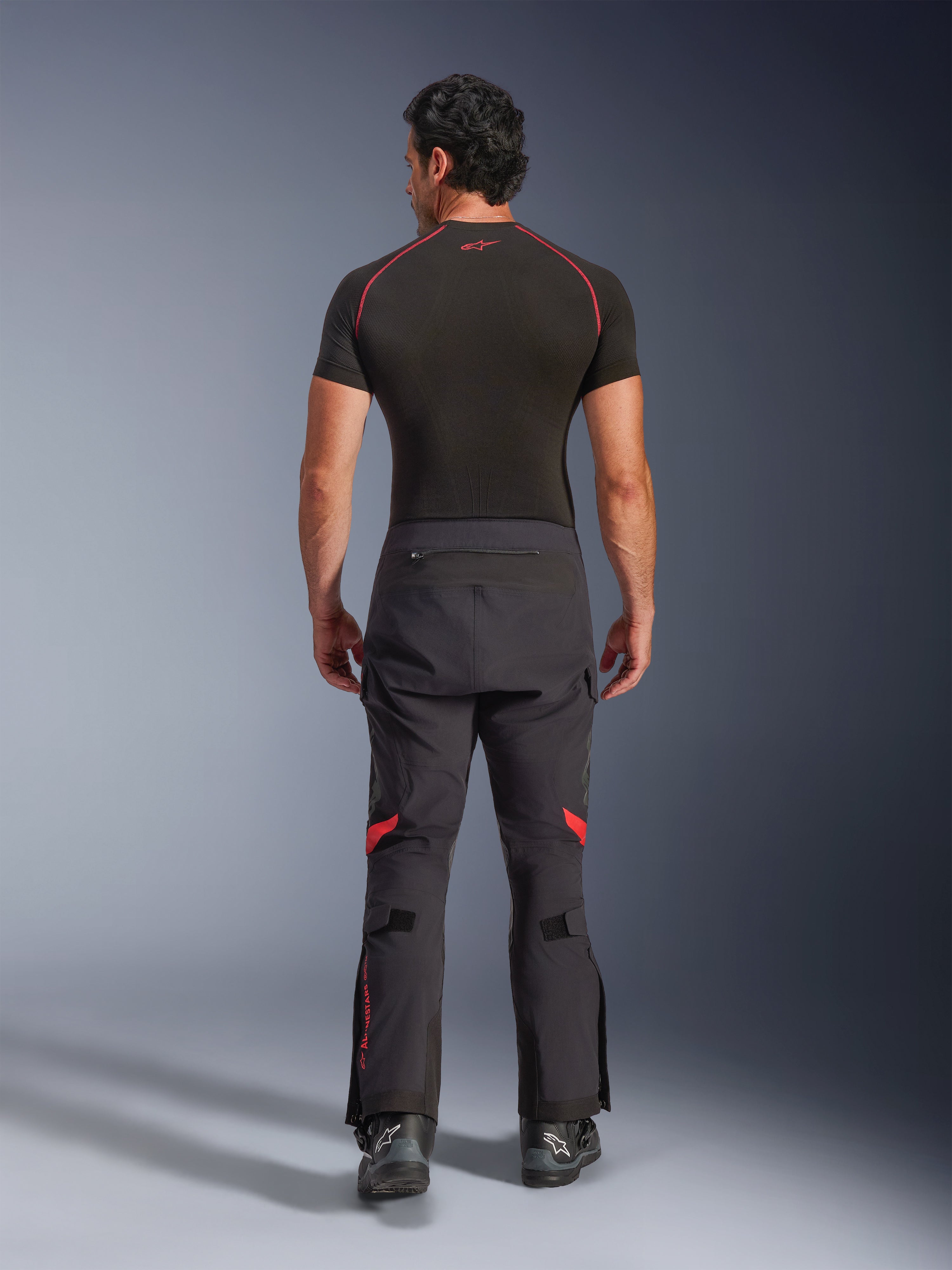 Alpinestars Halo Pro Drystar® XF laminerade byxor, äventyrsmotorcykelbyxor, Black Dark Shadow med röda detaljer, sedda bakifrån bärs med ett svart kortärmat underställ och svarta äventyrsstövlar, visar en hållbar 3-lagers laminerad konstruktion och ergonomisk passform.