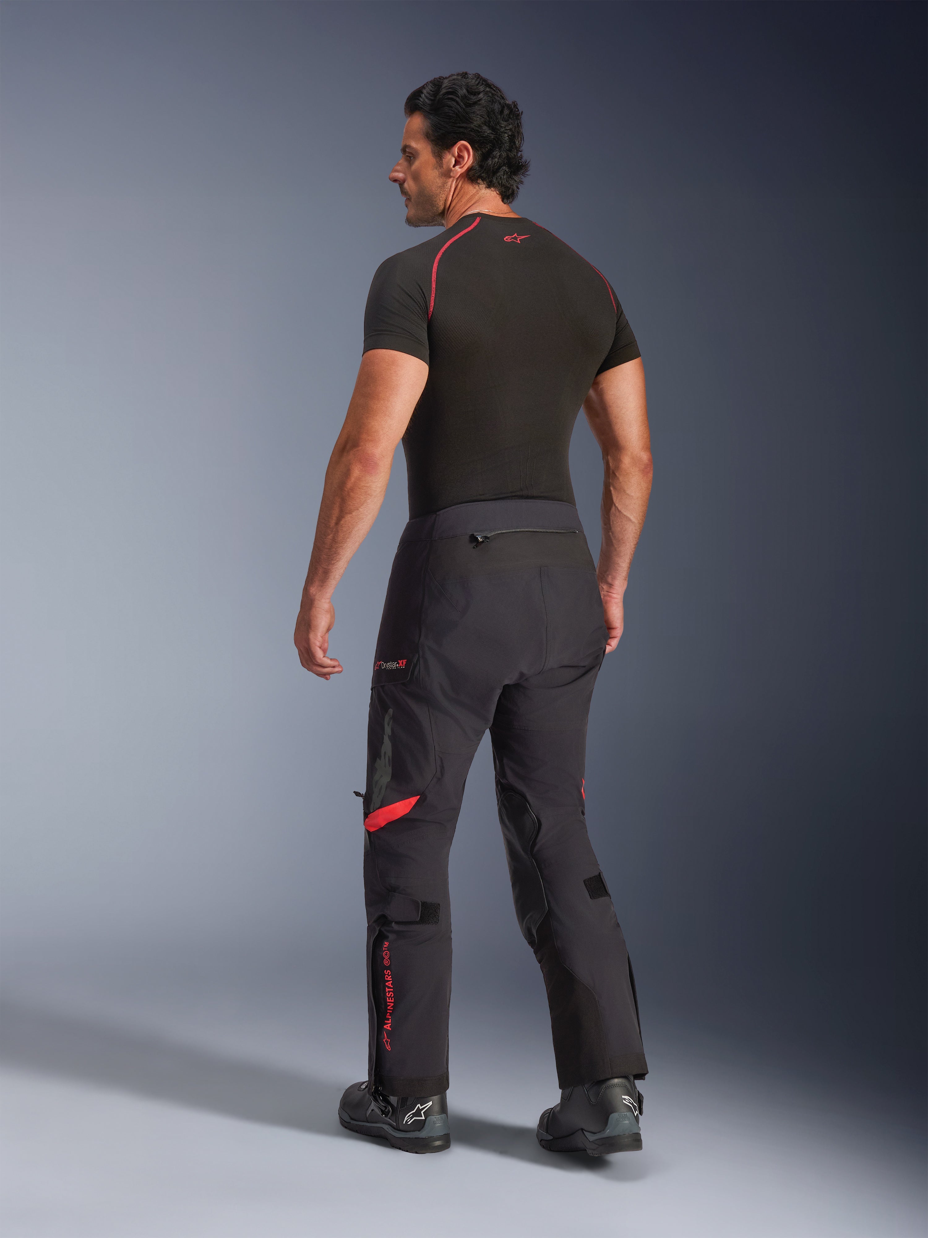 Alpinestars Halo Pro Drystar® XF laminerade byxor, Black Dark Shadow med röda detaljer, bakifrån på manlig modell, kombinerade med ett svart underställ och motorcykelstövlar, teknisk adventure-touringutrustning för terrängkörning
