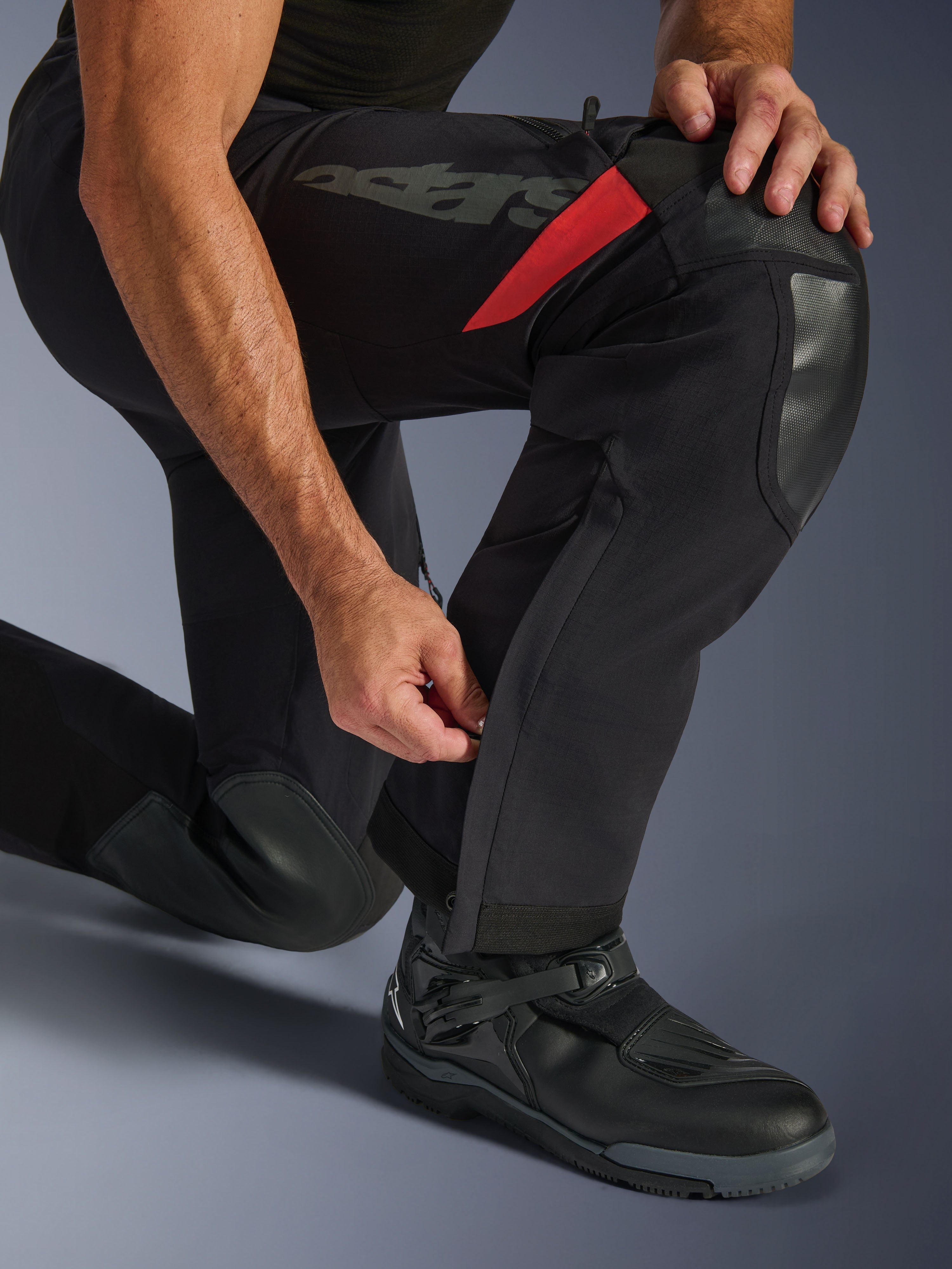 Alpinestars Halo Pro Drystar® XF laminerade byxor, Black Dark Shadow, närbild av underbenet med justerbar dragkedja vid vaden, med slitstarka knäförstärkningar, röda detaljer och 'astars'-logotyp, bärs med svarta motorcykelstövlar.