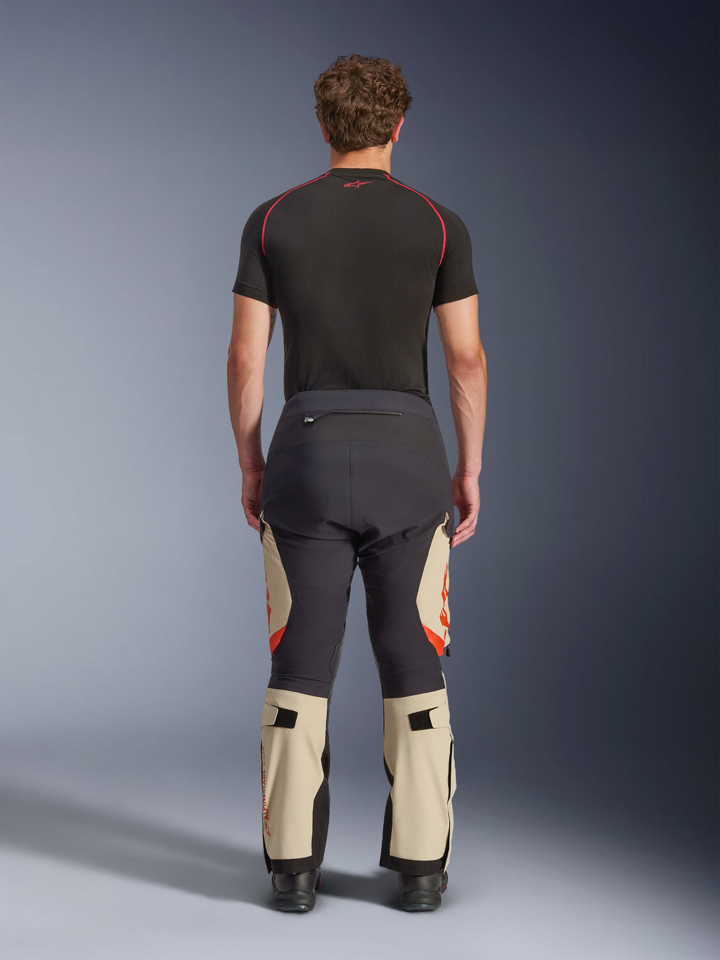Alpinestars Halo Pro Drystar® XF laminerade byxor, Mastic Black, sedda bakifrån på en modell, svart och beige färgblockering med orangea logotyper, äventyrsbyxor för motorcykel med hållbar textilkonstruktion och justerbara vadsektioner