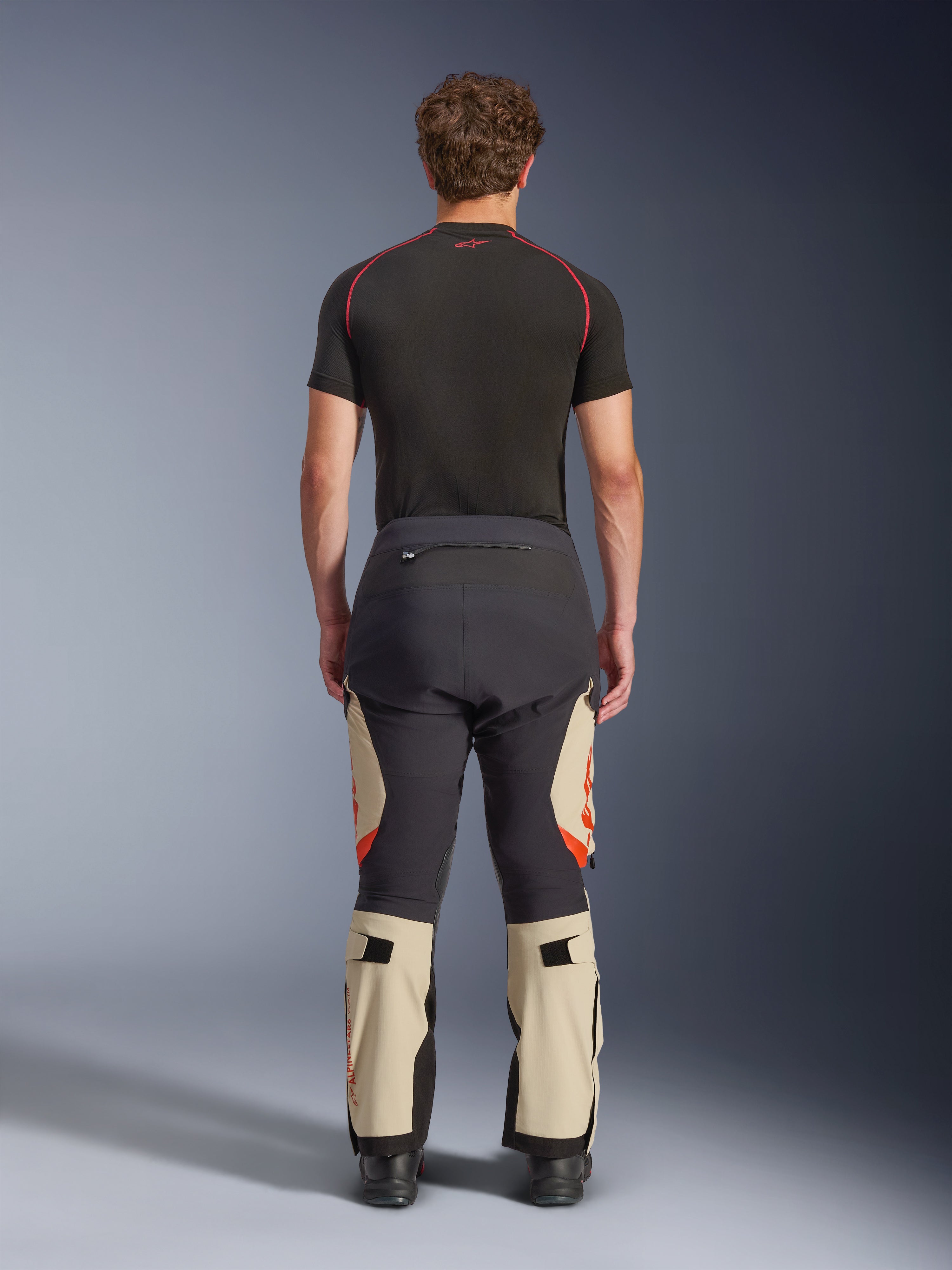 Alpinestars Halo Pro Drystar® XF laminerade byxor, Mastic Black, sedda bakifrån på en modell, svart och beige färgblockering med orangea logotyper, äventyrsbyxor för motorcykel med hållbar textilkonstruktion och justerbara vadsektioner