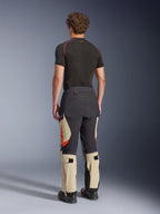 Alpinestars Halo Pro Drystar® XF Laminerade Byxor, äventyrsbyxor för motorcykel, Mastic Black och beige med orange detaljer, vy bakifrån som visar 3-lagers laminerad skalkonstruktion, kombinerad med ett svart underställ och äventyrsstövlar.