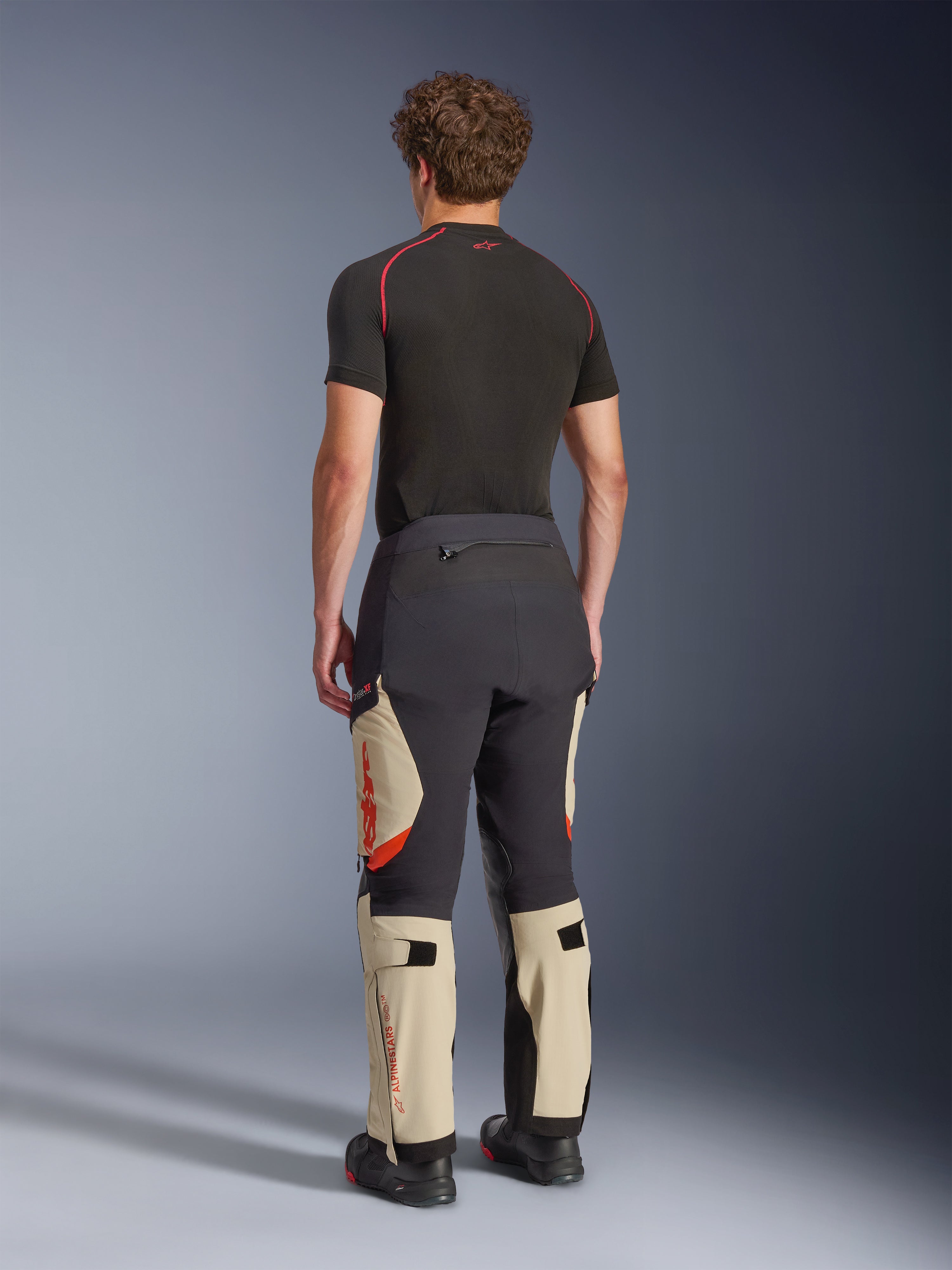 Alpinestars Halo Pro Drystar® XF Laminerade Byxor, äventyrsbyxor för motorcykel, Mastic Black och beige med orange detaljer, vy bakifrån som visar 3-lagers laminerad skalkonstruktion, kombinerad med ett svart underställ och äventyrsstövlar.