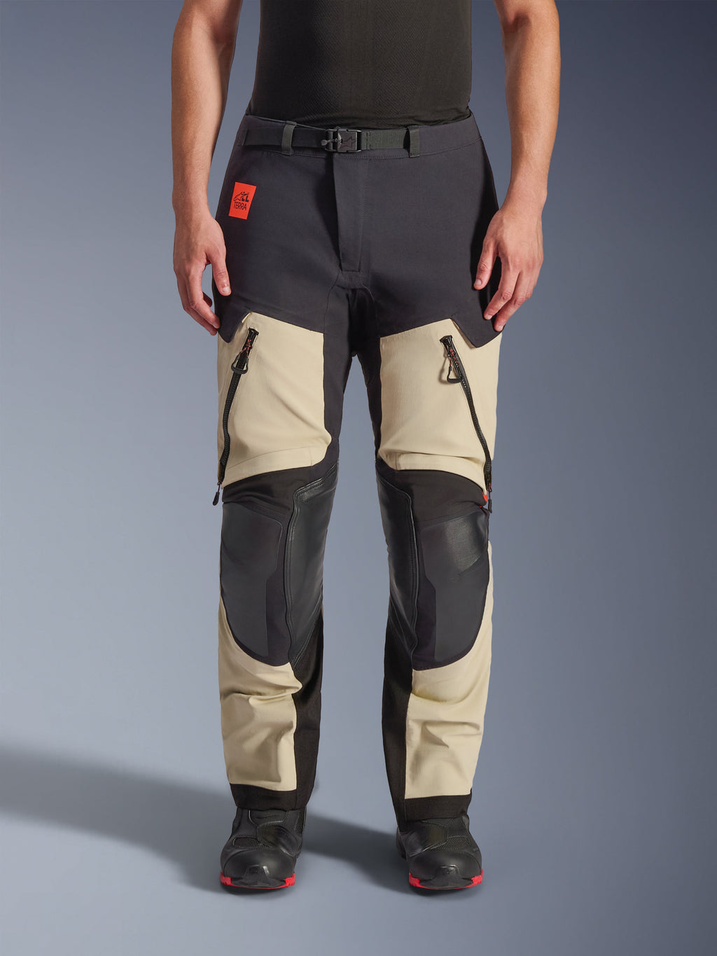 Alpinestars Halo Pro Drystar® XF laminerade byxor, äventyrsbyxor för motorcykel, Mastic Black, frontvy buren av en förare, med stora lårventiler med dragkedja, förstärkta svarta knäpaneler och beige textilkonstruktion, kombinerade med svarta och röda mc-stövlar.