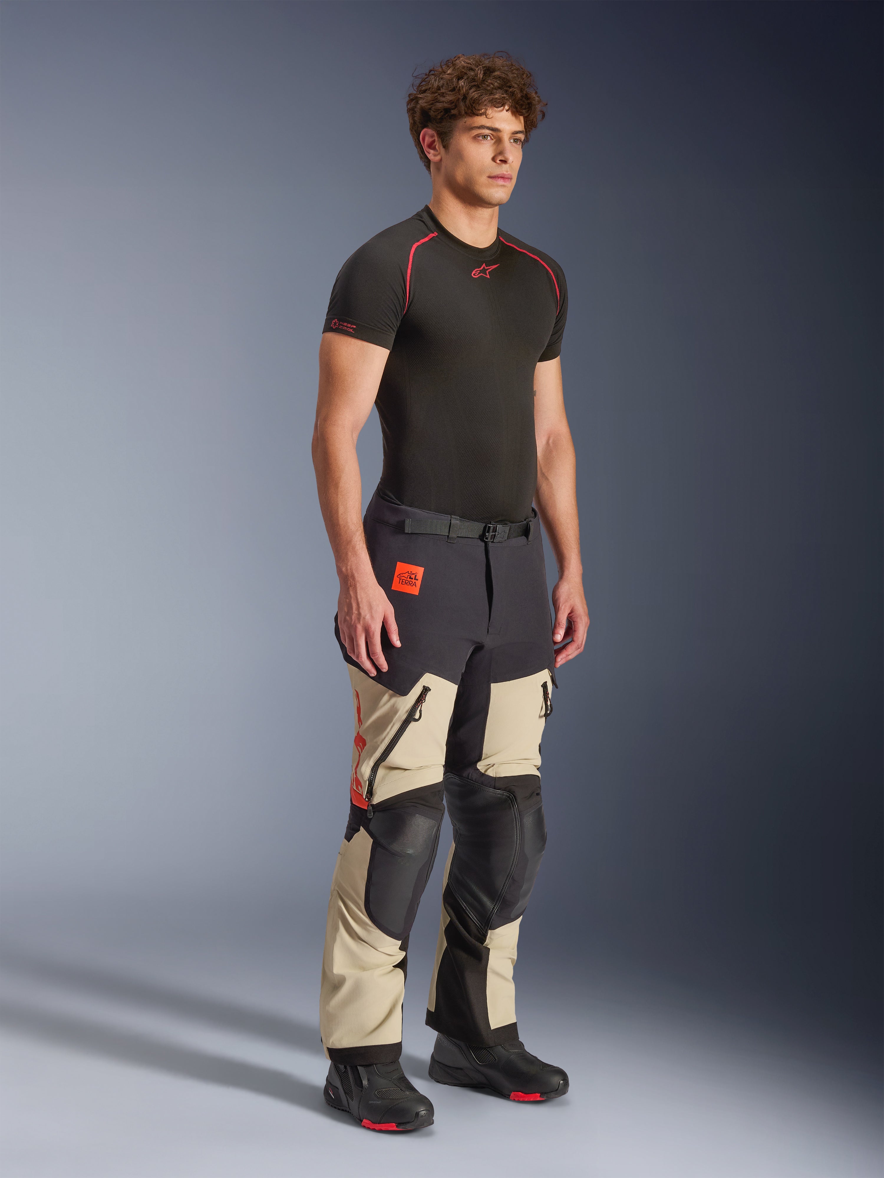 Alpinestars Halo Pro Drystar® XF laminerade byxor i Mastic Black, äventyrsmotorcykelbyxor som bärs av en modell, med svarta knä- och sätesförstärkningar, orange logotypdetaljer och flera funktionella dragkedjor, stylade med ett svart underställ och mc-stövlar.