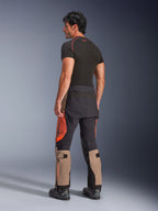 Alpinestars Halo Pro Drystar® XF laminerade byxor i Walnut Rust Black, vy bakifrån av en manlig modell som bär äventyrsmotorcykelbyxor med beige, orangea och svarta paneler, stylad med ett svart kortärmat underställ och svarta motorcykelstövlar, framhäver teknisk utrustning för äventyrskörning