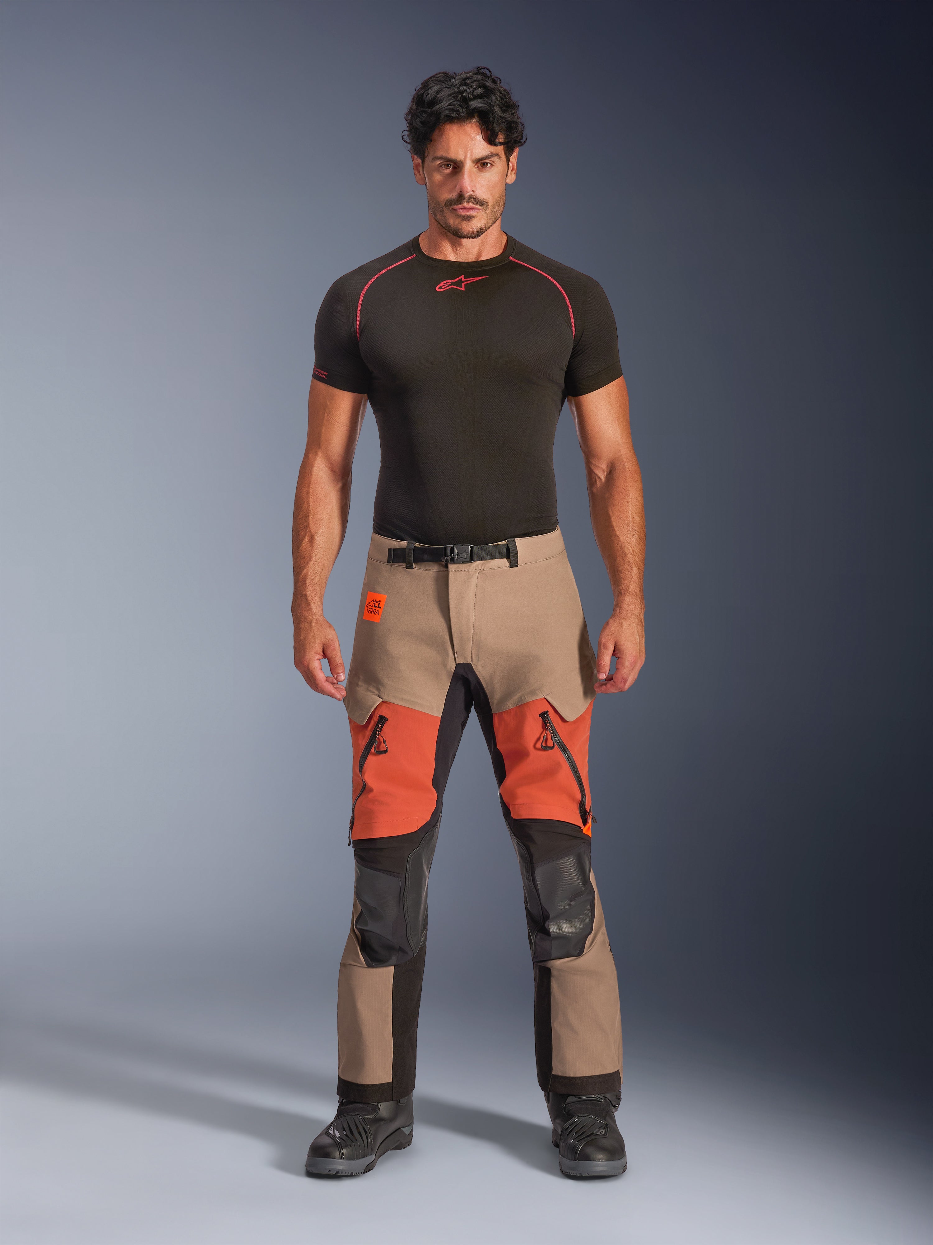Manlig modell iförd Alpinestars Halo Pro Drystar® XF laminerade byxor, äventyrsmotorcykelbyxor, Walnut Rust Black, beige och orange färgblockad design med svarta tekniska paneler, lårventiler med dragkedja och magnetiskt midjespänne, kombinerat med ett svart baslager och svarta motorcykelstövlar