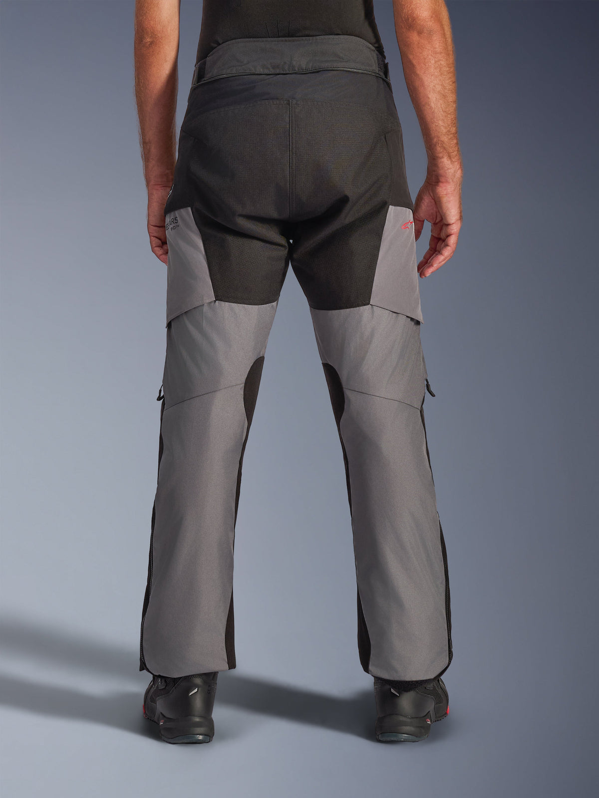 Alpinestars Andes V4 Drystar® byxor, adventure-motorcykelbyxor, svarta och mörkgrå, vy bakifrån som visar förstärkt svart säte och grå ben, med en liten röd logotyp, kombinerade med svarta mc-stövlar, slitstark textilkonstruktion för touring i alla väder.